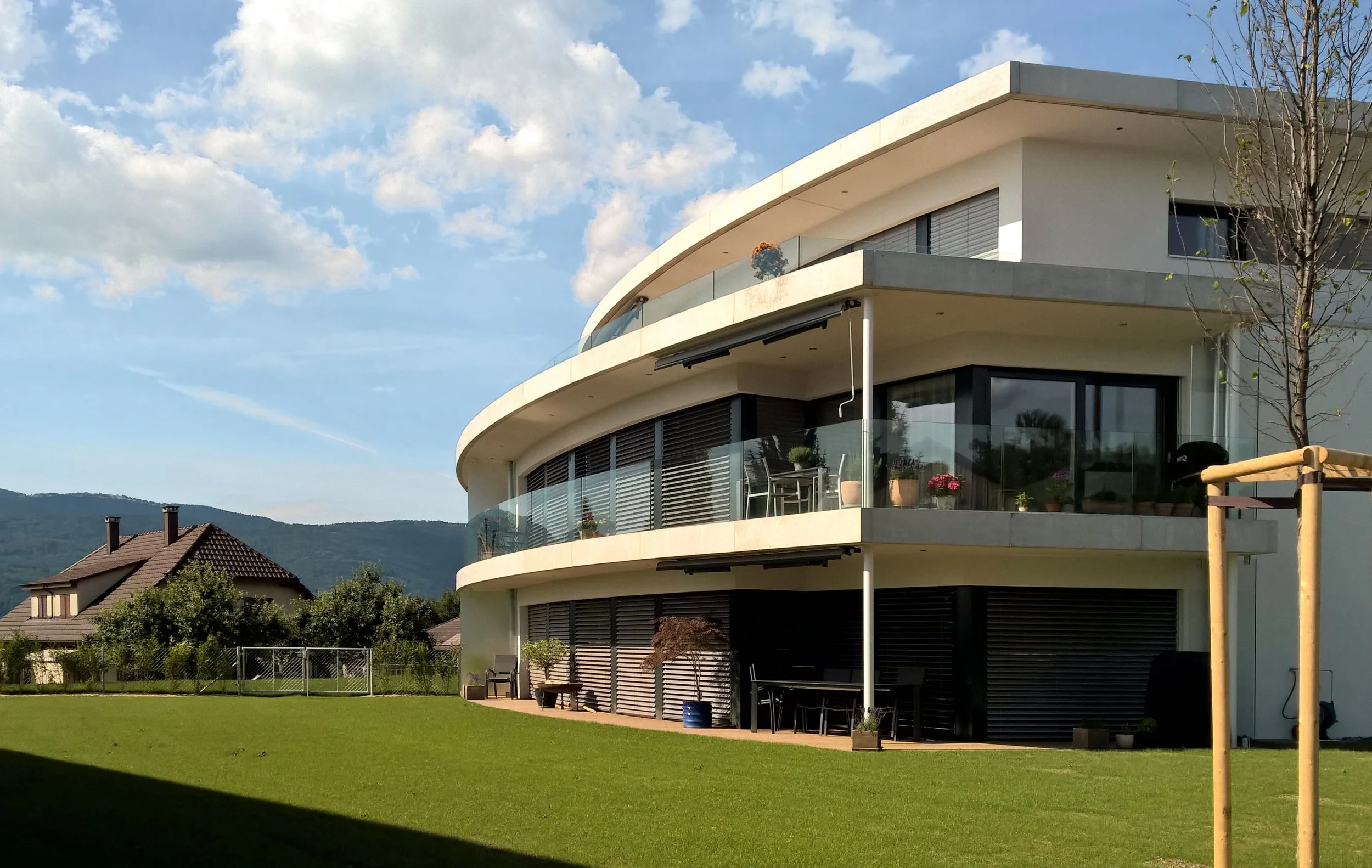 Modernes, mehrstöckiges Gebäude mit großen Balkonen und Glasgeländern, umgeben von einem grünen Rasen und Bäumen, bei sonnigem Himmel.