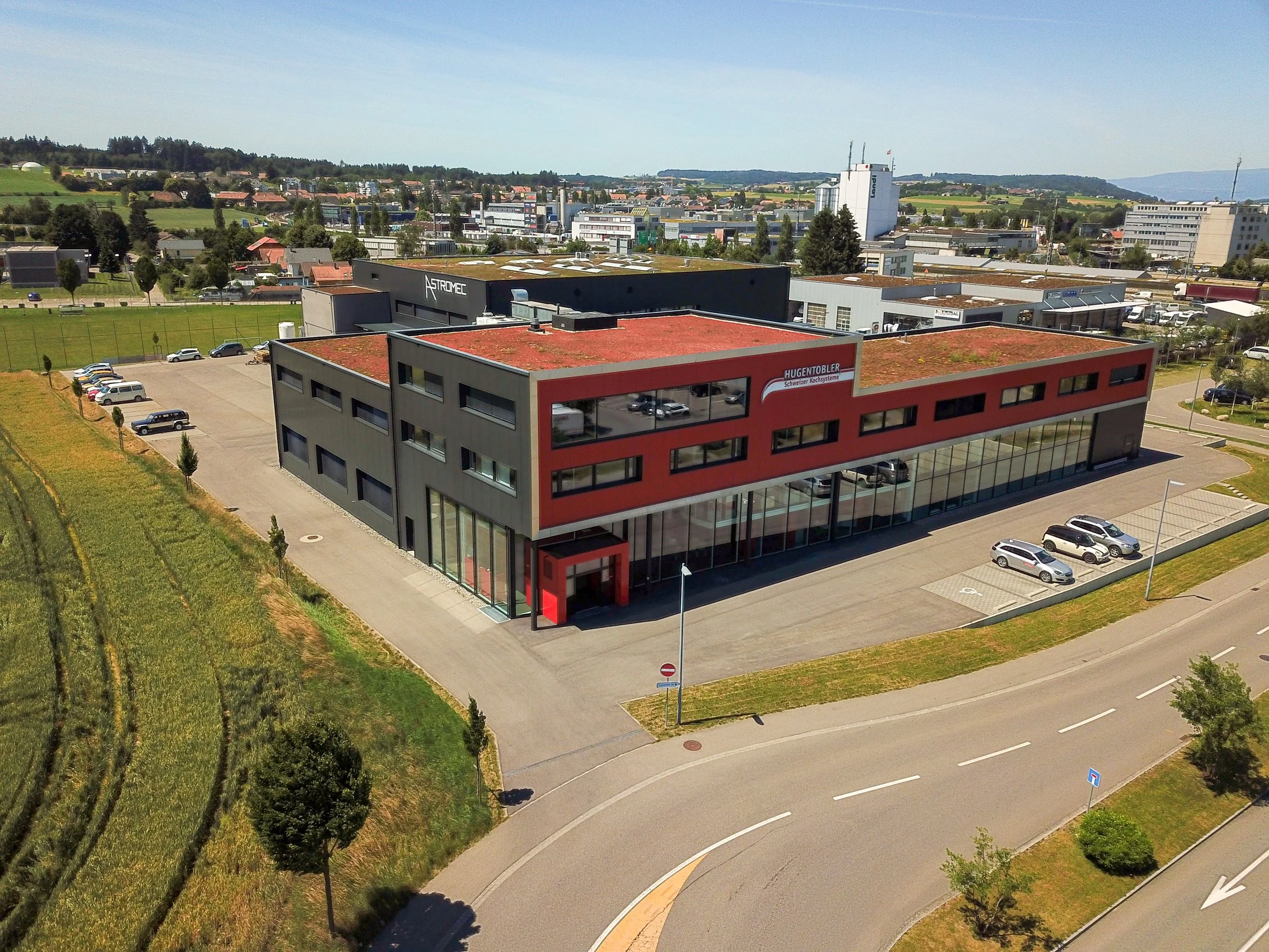 Luftaufnahme eines modernen Bürogebäudes mit roten und grauen Fassaden, umgeben von Parkplätzen, in einer Stadt mit grünen Hügeln im Hintergrund.