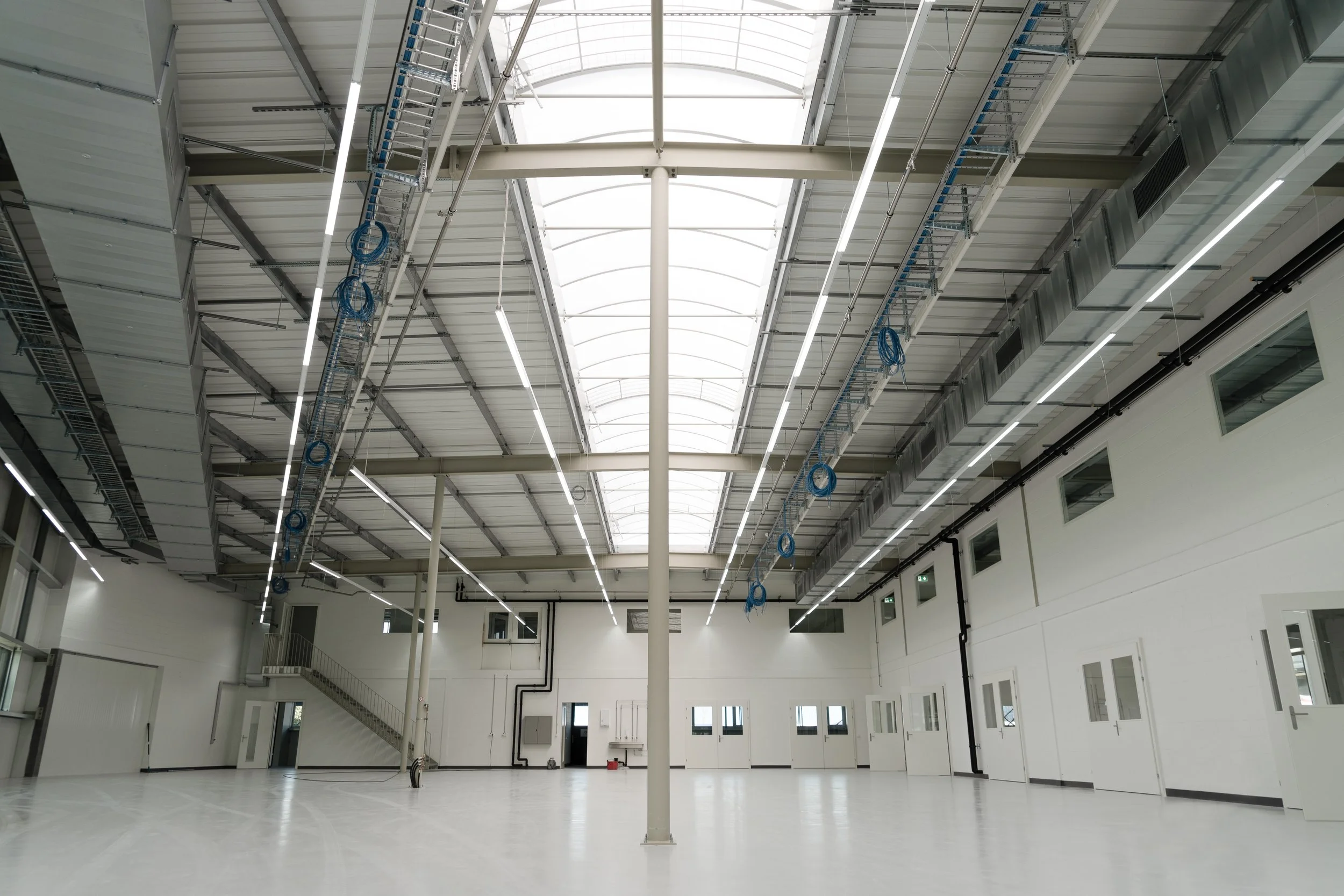 Großer, heller Raum mit hoher Decke und vielen Fenstern, in einer modernen Fabrik oder Lagerhalle, mit Kabeln und Leitern an der Decke installiert.