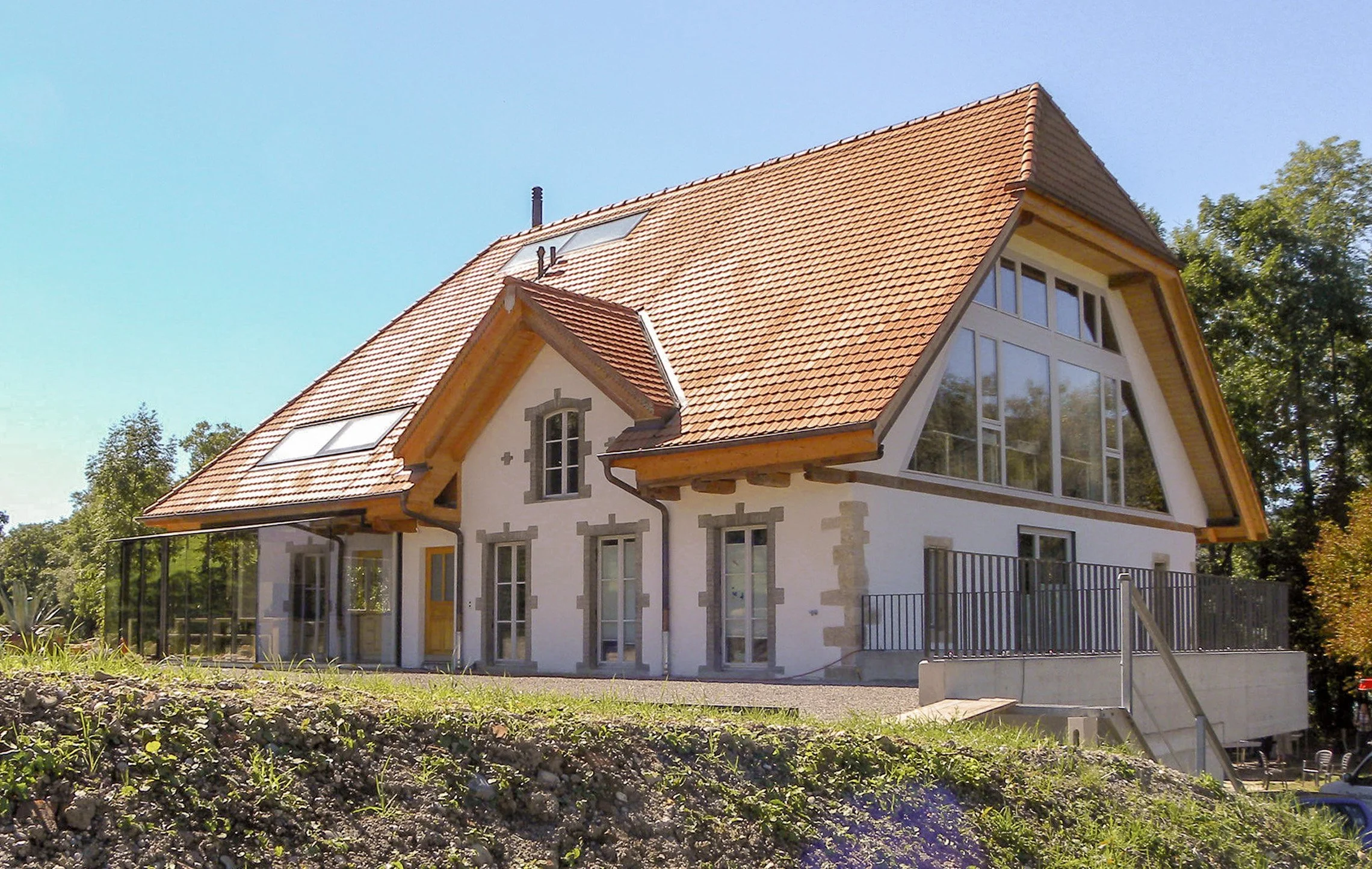 Modernes Mehrfamilienhaus mit roten Dachziegeln, großen Fenstern und einem Balkon, umgeben von Bäumen und Garten.