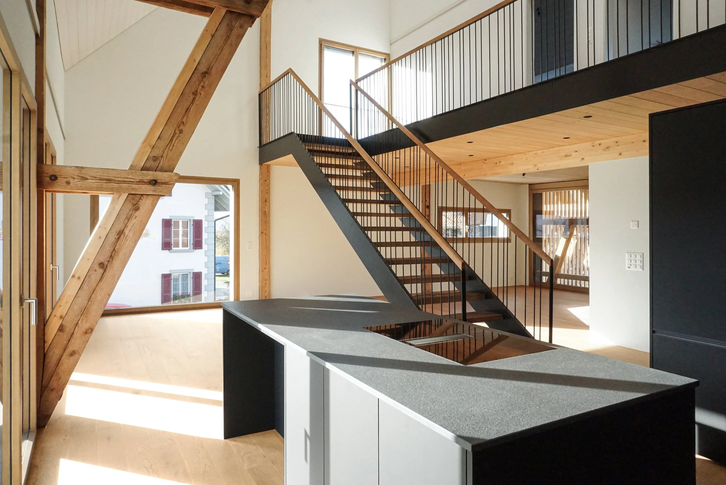 Innenansicht eines modernen Hauses mit Holz und schwarzen Akzenten, sichtbare offene Treppe, große Fenster, viel natürliches Licht, Klimaanlage an der Wand, Holzboden.
