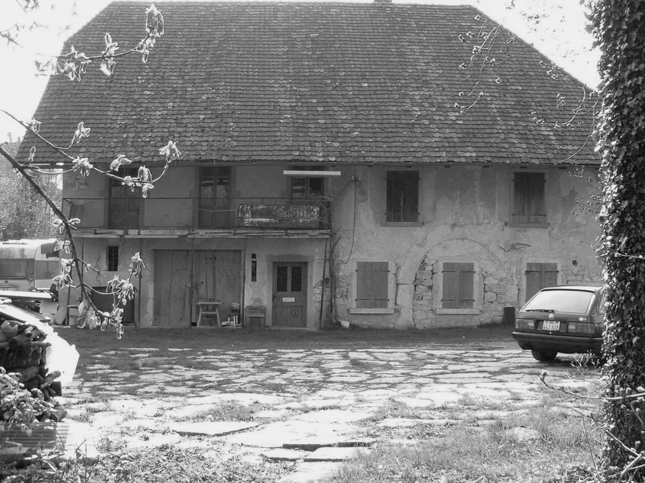 Ein altes, zweistöckiges Haus mit einer großen, schieferbedeckten Dach, außenliegenden Balkonen und verschlossenen Fensterläden. Im Vordergrund befinden sich ein geparkter Wagen, ein alter Holzstuhl und eine Kiste. Der Garten wirkt vernachlässigt. Das Bild ist in Schwarzweiß gehalten.