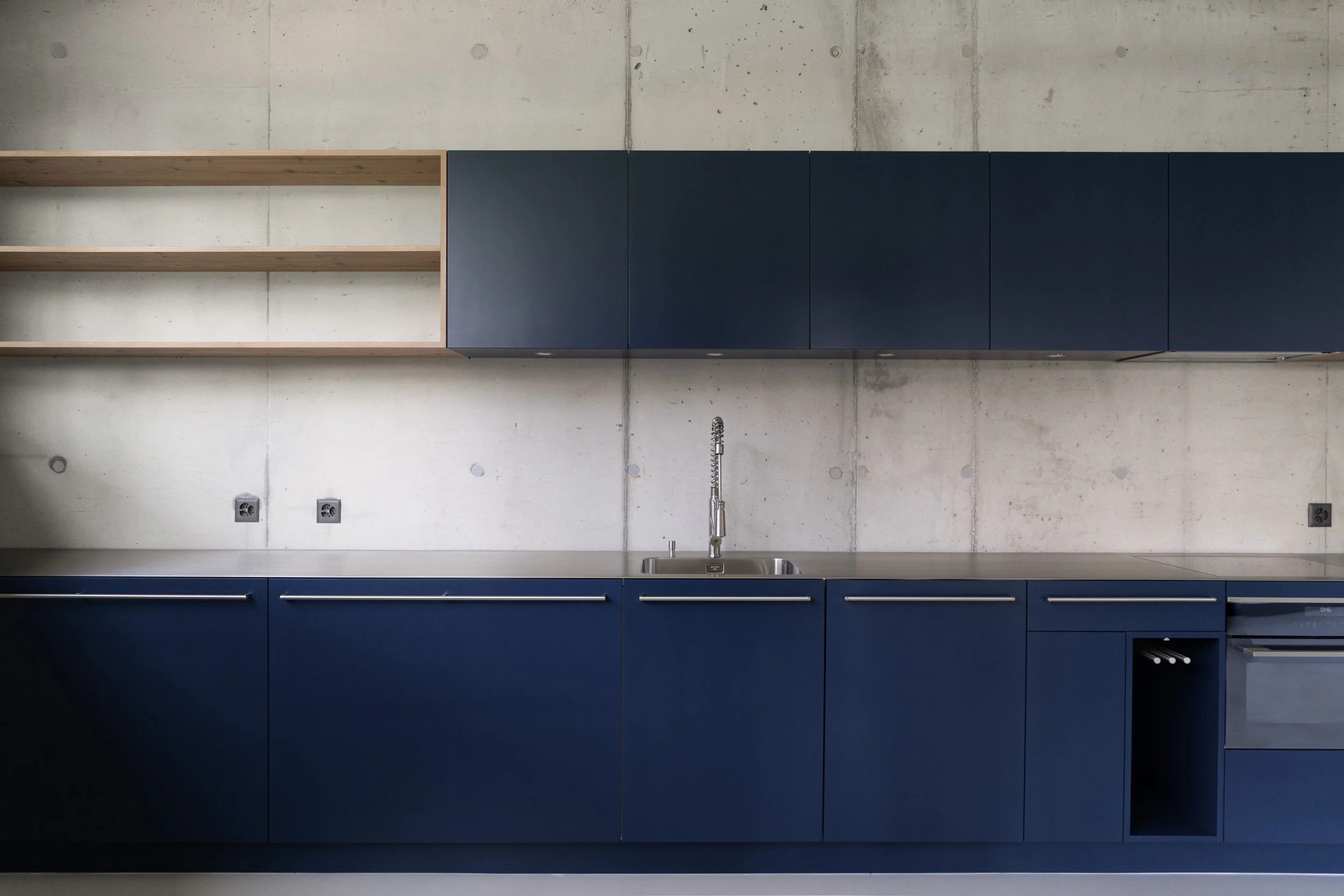 Moderne Küchenzeile mit blauen Schränken, einem Edelstahlspülbecken, schwarzen Elektroanschlüssen und offenen Regalen gegen eine Betonwand.