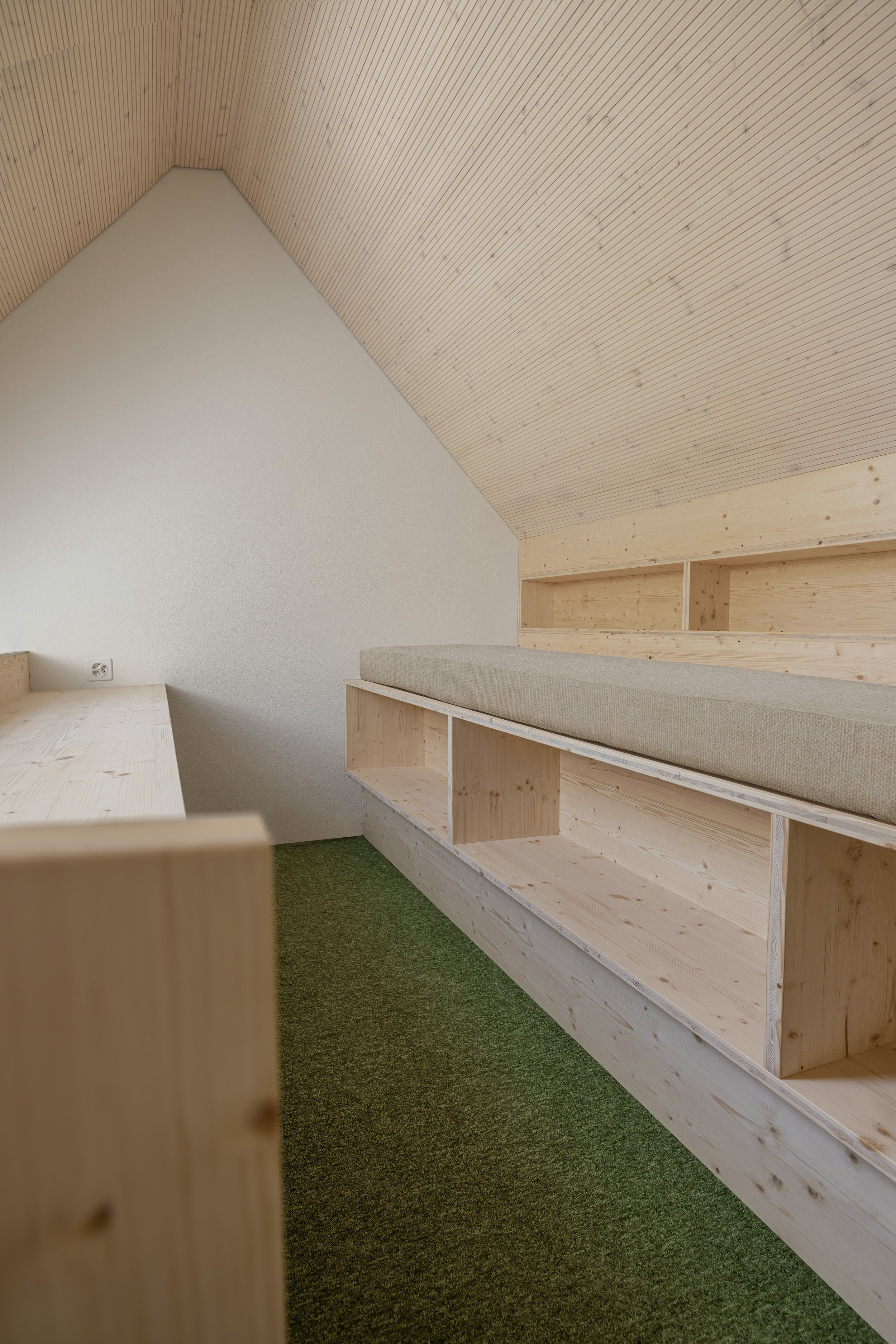 Innenaufnahme eines Raumes mit Holzdecke, weißer Wand und grüner Teppichboden, dazu eine offene Holzregalwand.