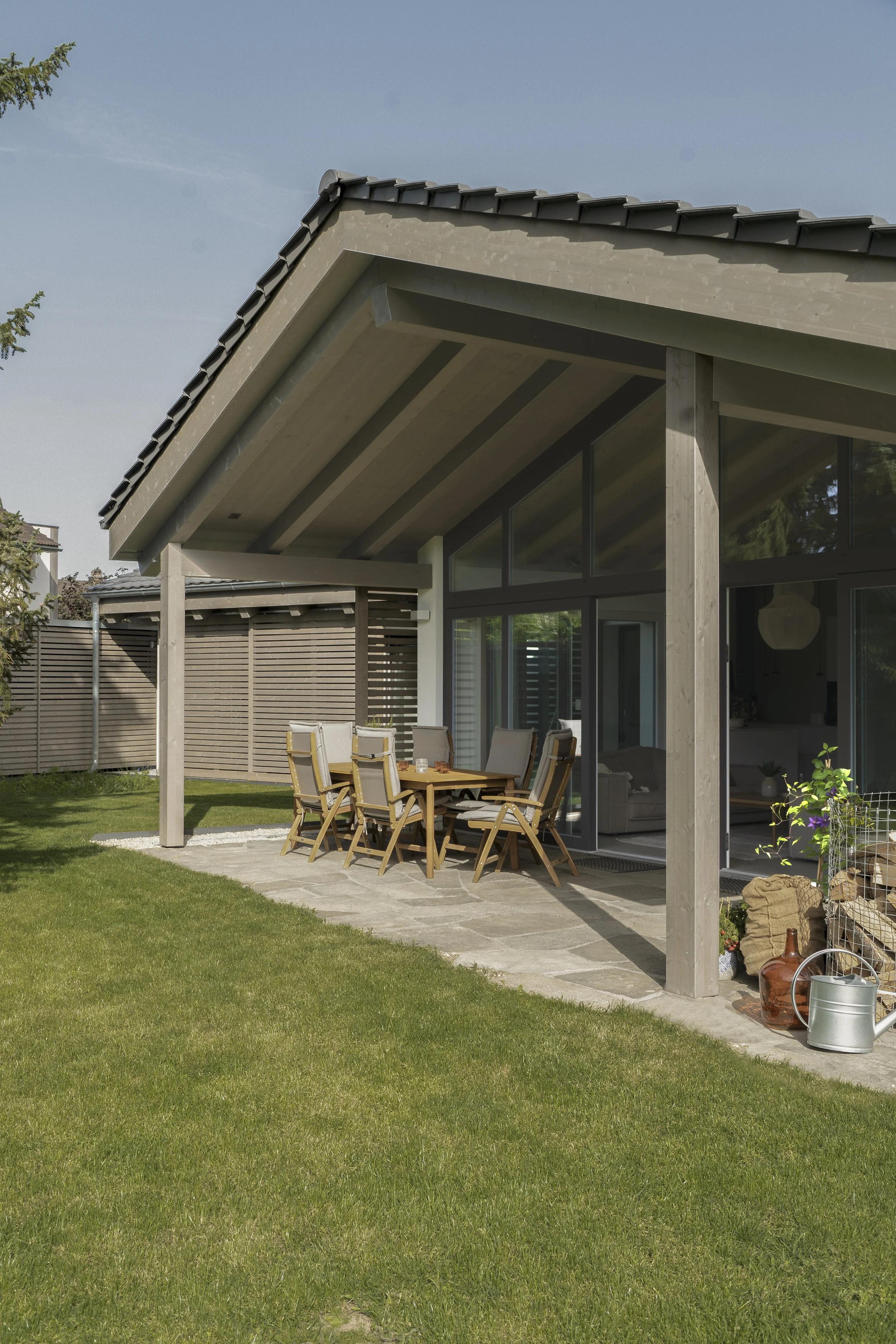 Moderne Terrasse mit Gartentisch und Stühlen vor einem Haus mit großen Fenstern und einem überdachten Bereich, im Garten sind Gartenwerkzeuge und Dekorationen zu sehen, umgeben von Grünflächen.