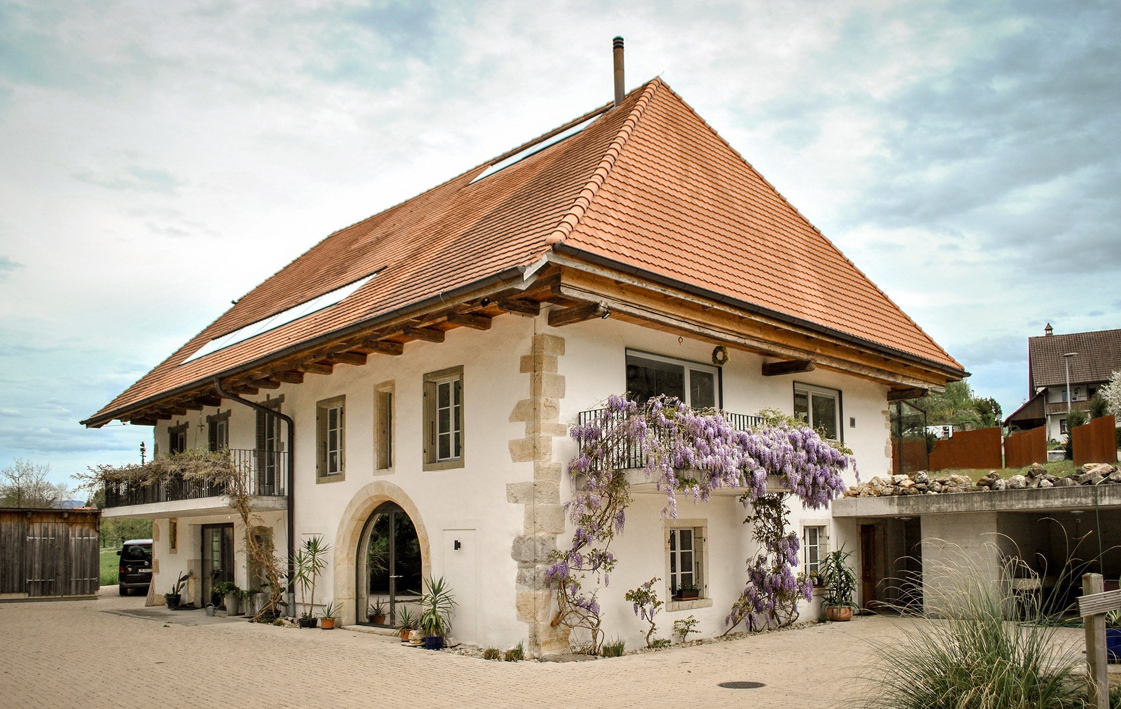 Ein traditionelles Haus mit roten Ziegeldächern, weißen Wänden und blühenden Pflanzen im Garten. Das Haus hat mehrere Fenster und einen Balkon mit einer Blumenkletterpflanze.