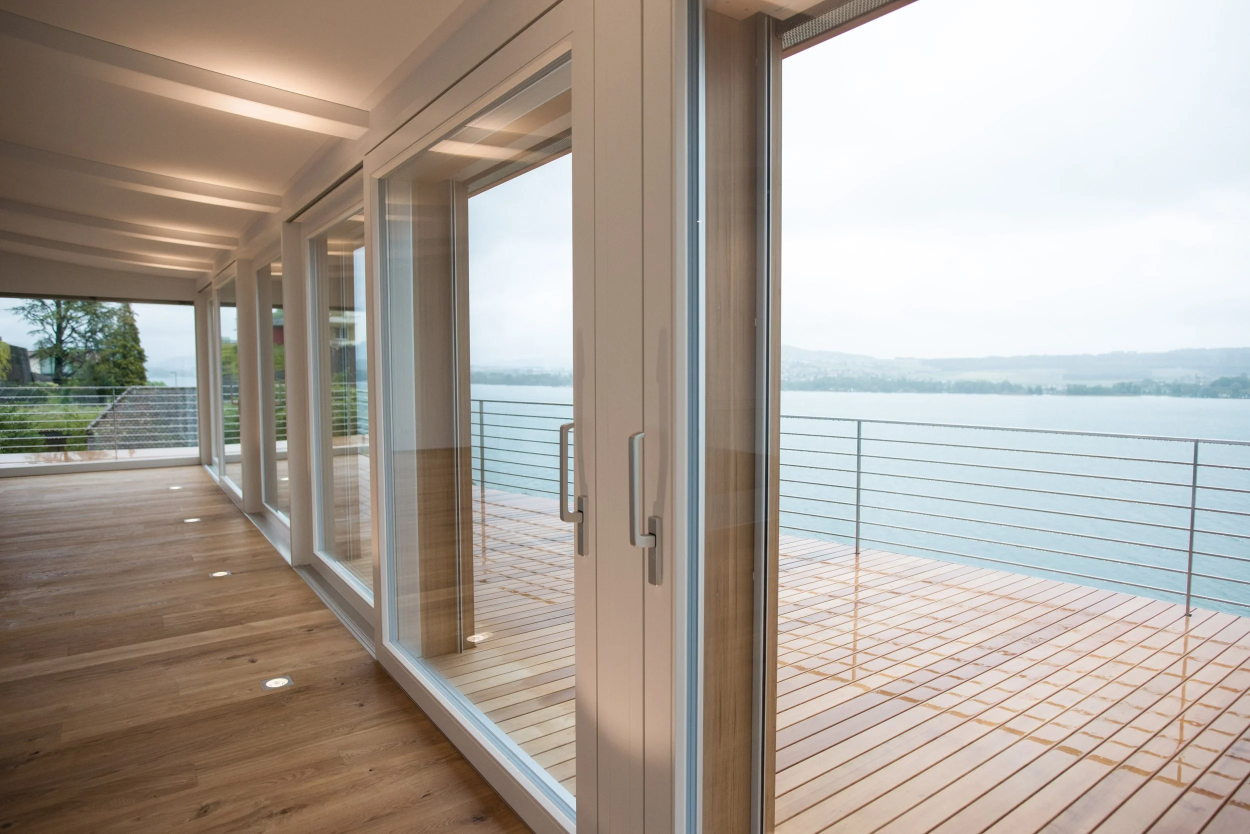 Moderne Wohnraum mit großen Glas-Terrassentüren, Holzboden und Blick auf eine Wasserlandschaft im Hintergrund.