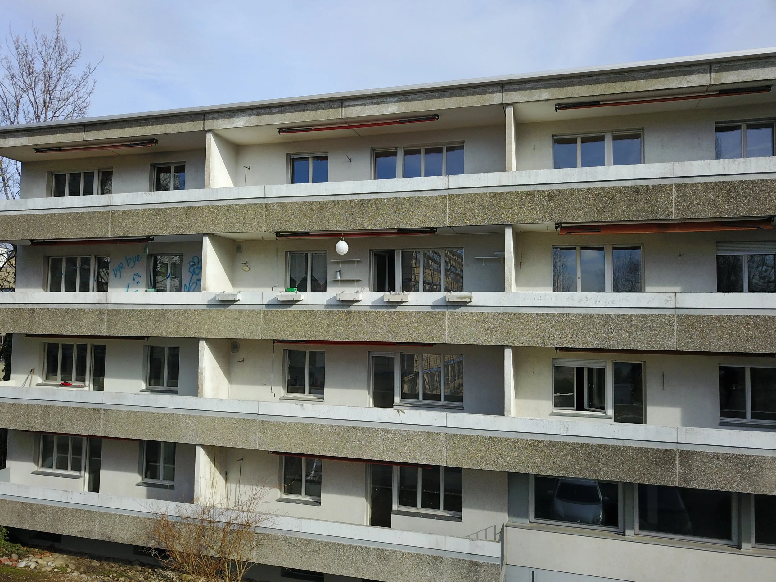 Mehrstöckiges modernes Wohngebäude mit Balkonen und großen Fenstern, teilweise mit dekorativen Elementen, im Freien mit einigen Bäumen und einem kleinen Garten