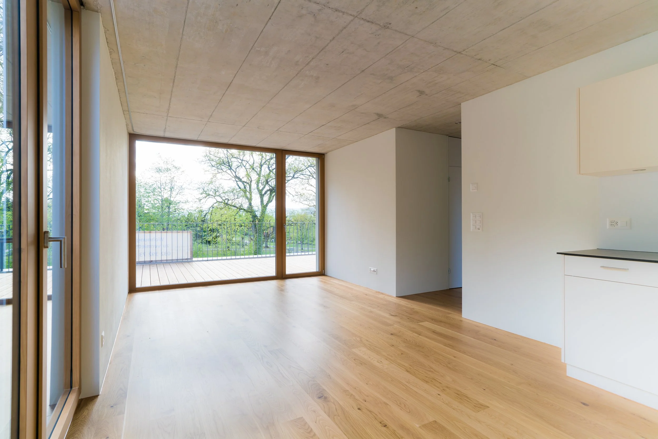 Innenraum eines modernen, hellen Zimmers mit großen Fenstern und Holzboden, Blick nach draußen auf Bäume und eine Terrasse.