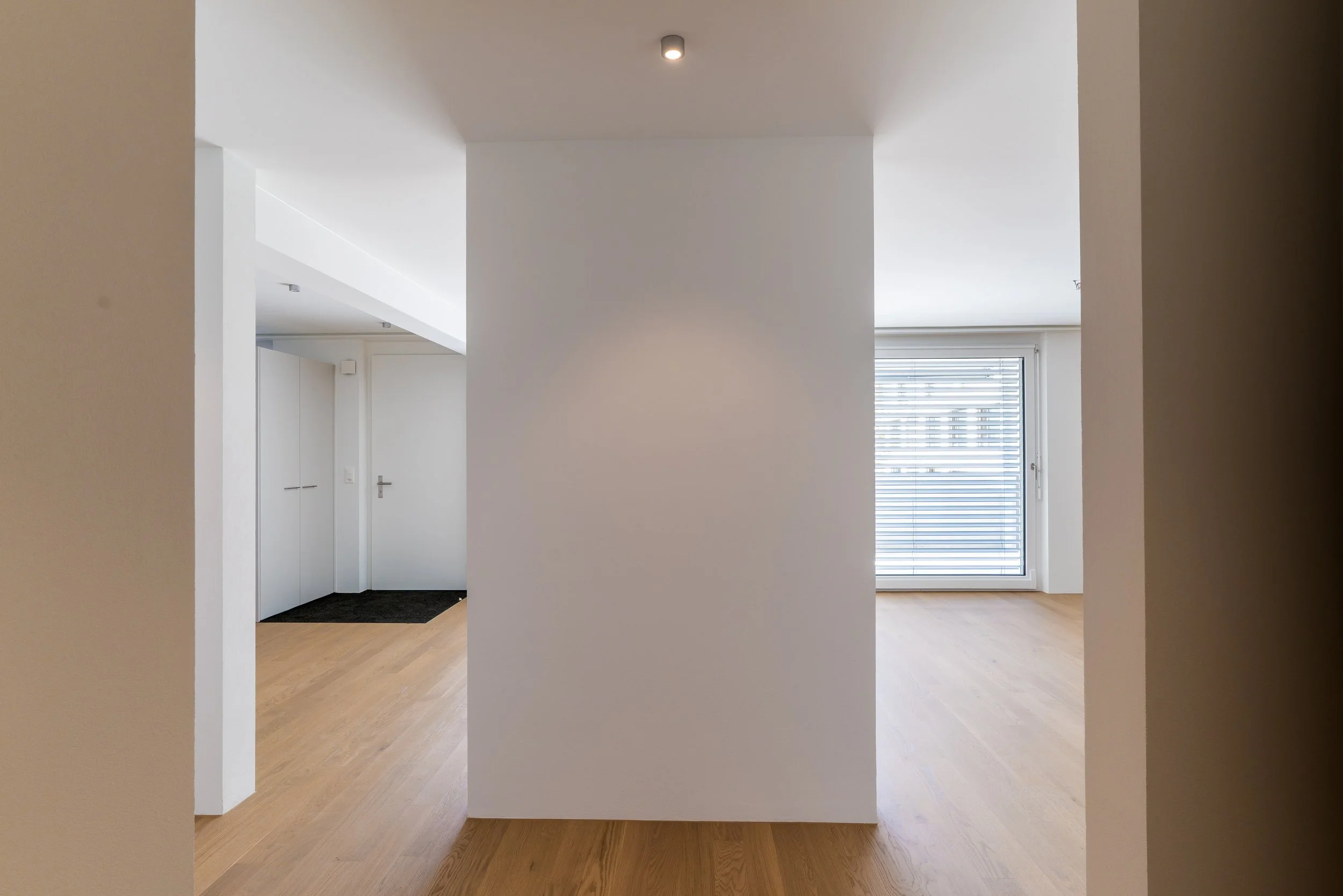 Ein minimalistisch eingerichteter Raum mit hellem Holzboden und weißen Wänden. Im Hintergrund sind Fenster mit horizontalen Jalousien sichtbar, die Tageslicht hereinlassen. Im linken Bereich befindet sich eine weiße Tür, und der Raum wirkt modern und schlicht.