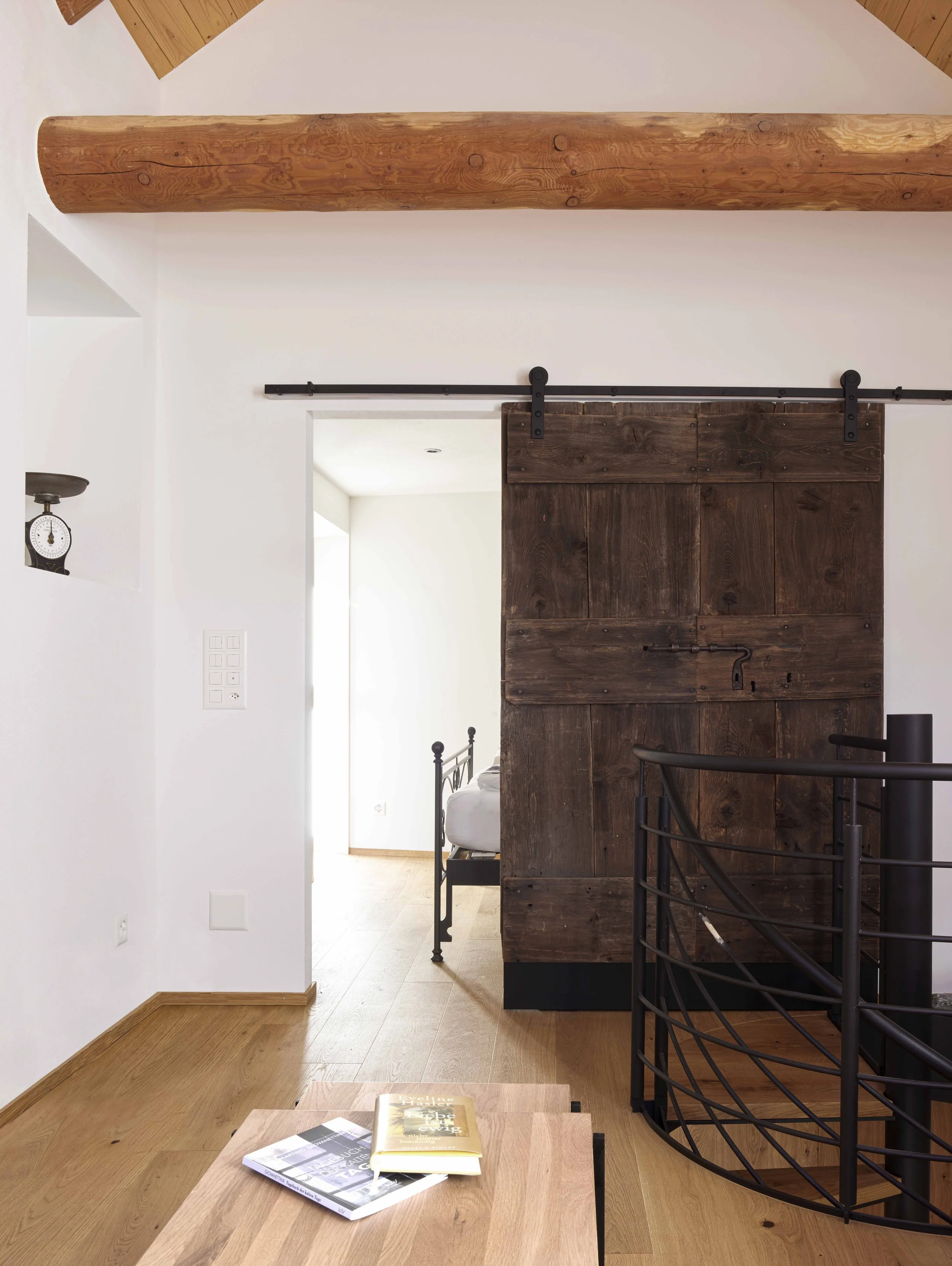 Innenraum mit Holz- und Metallmöbeln, weißen Wänden und einer Treppe, Blick auf ein Schlafzimmer im Hintergrund, Zeitschriften auf einem Tisch im Vordergrund.