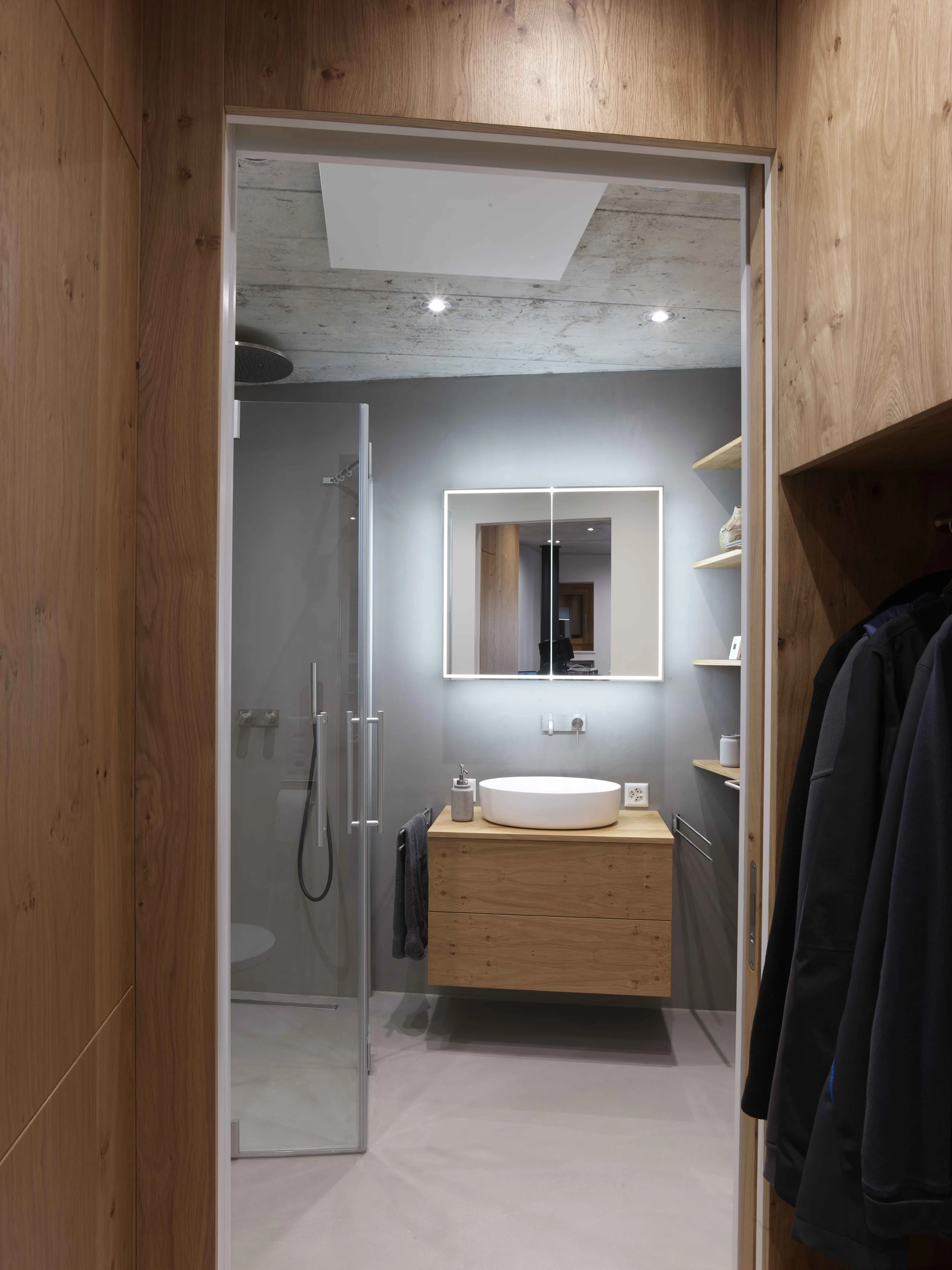 Ein moderner Waschraum mit einer Holz-Waschtisch, einer ovalen weißen Waschschale, einem großen Spiegel, grauem Wandanstrich, einer offenen Holzregal und Garderobe mit Dunklen Mänteln.