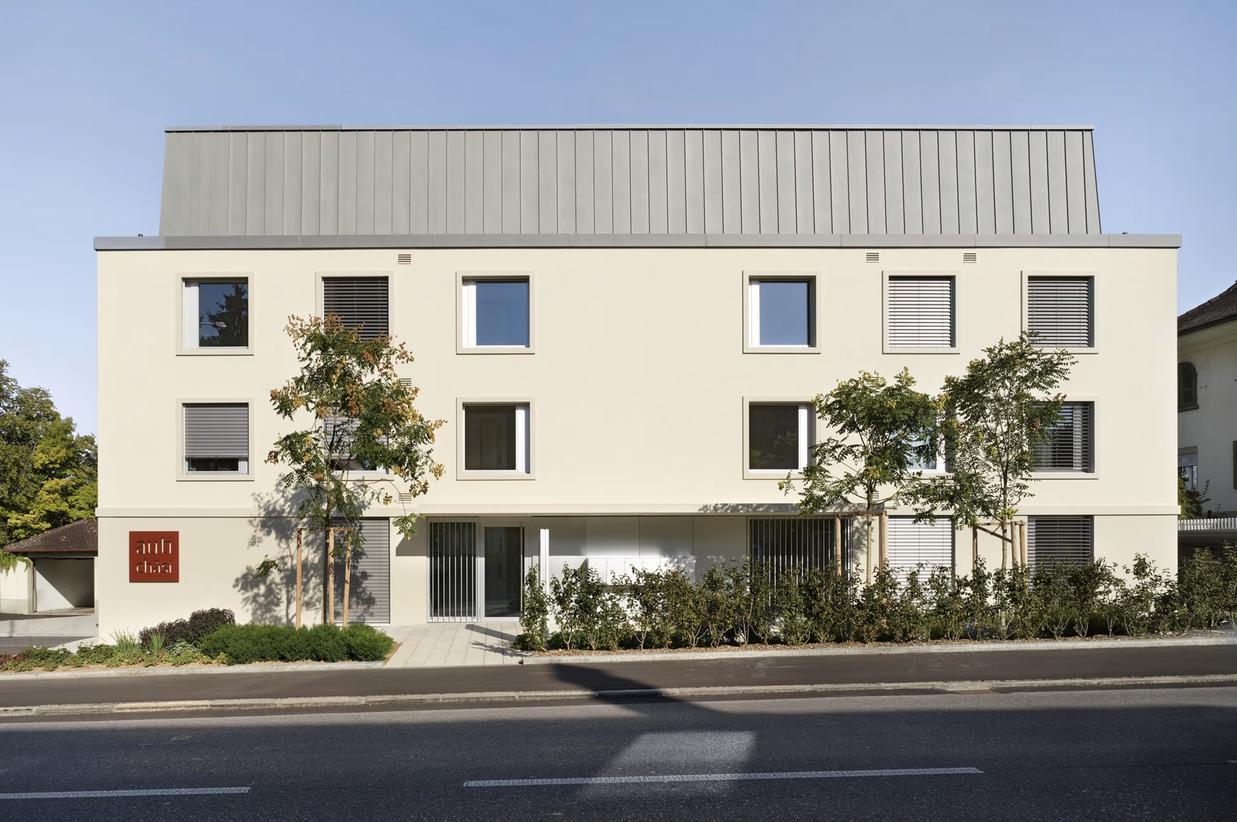 Moderne weiße Mehrfamilienhaus mit wenigen Fenstern und umgeben von kleinen Bäumen, vor einer Straße in einer städtischen Gegend bei sonnigem Himmel.