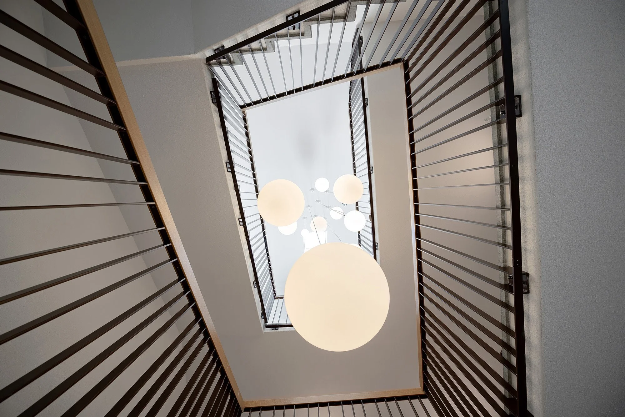 Blick nach oben auf eine Treppe mit Metallgeländer und runden Deckenleuchten.
