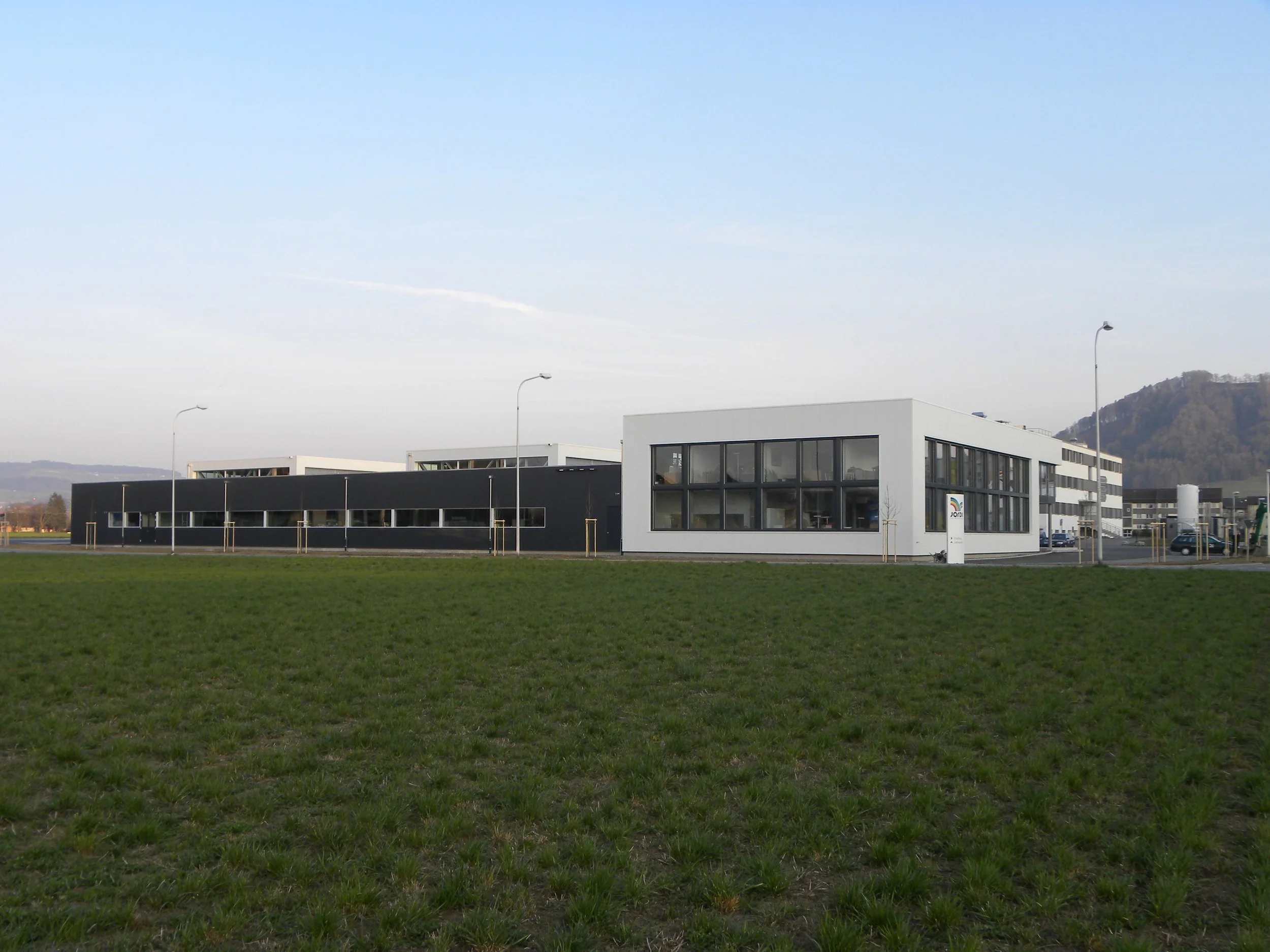 Moderne schwarze und weiße Bürogebäude mit großen Fenstern, umgeben von einem grünen Rasen und Straßenlaternen, im Hintergrund Berge und Himmel.