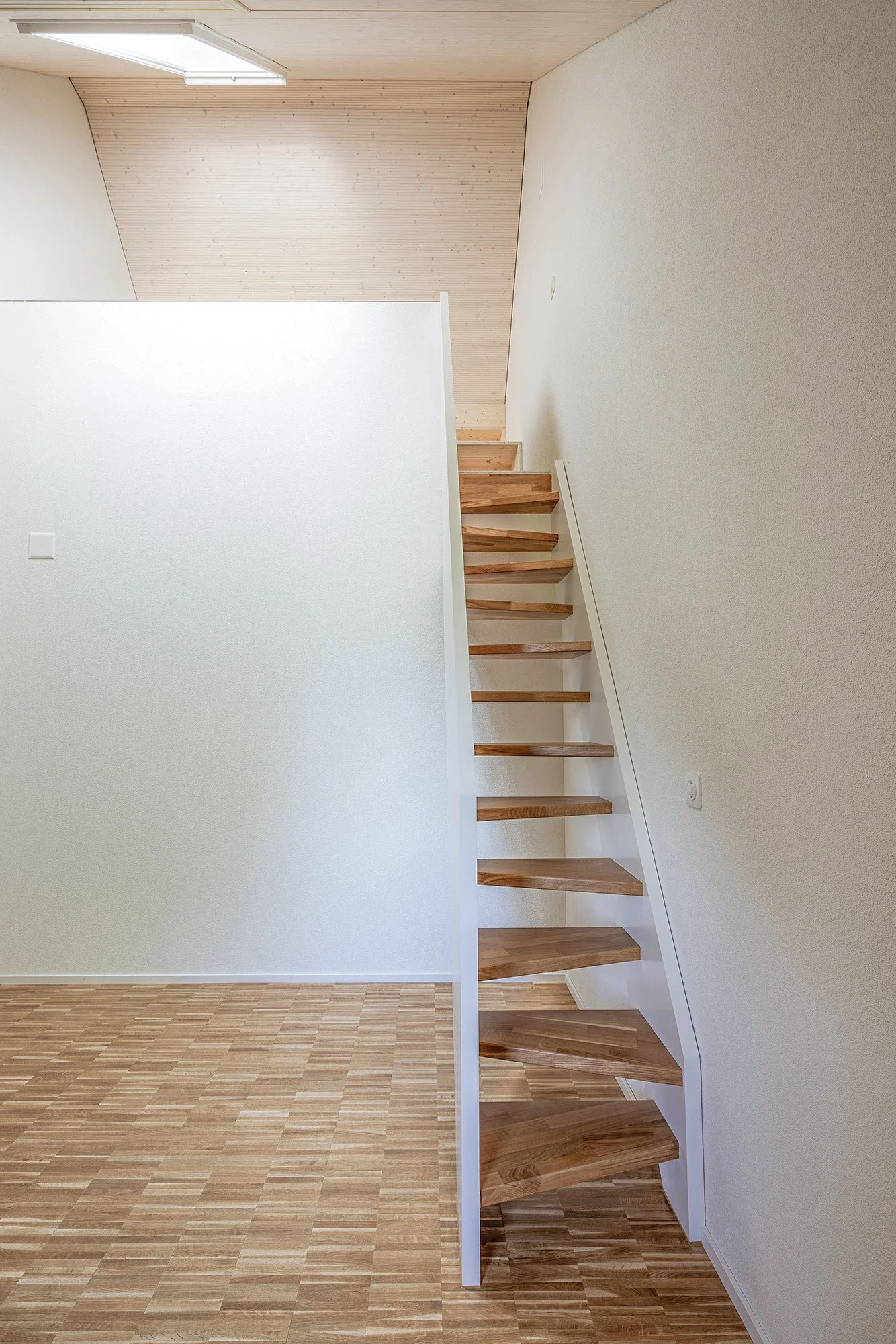 Innenaufnahme eines Hauses mit einer Treppe und Holzfußboden, weißen Wänden und einer Holzdecke unter einem Dachfenster.
