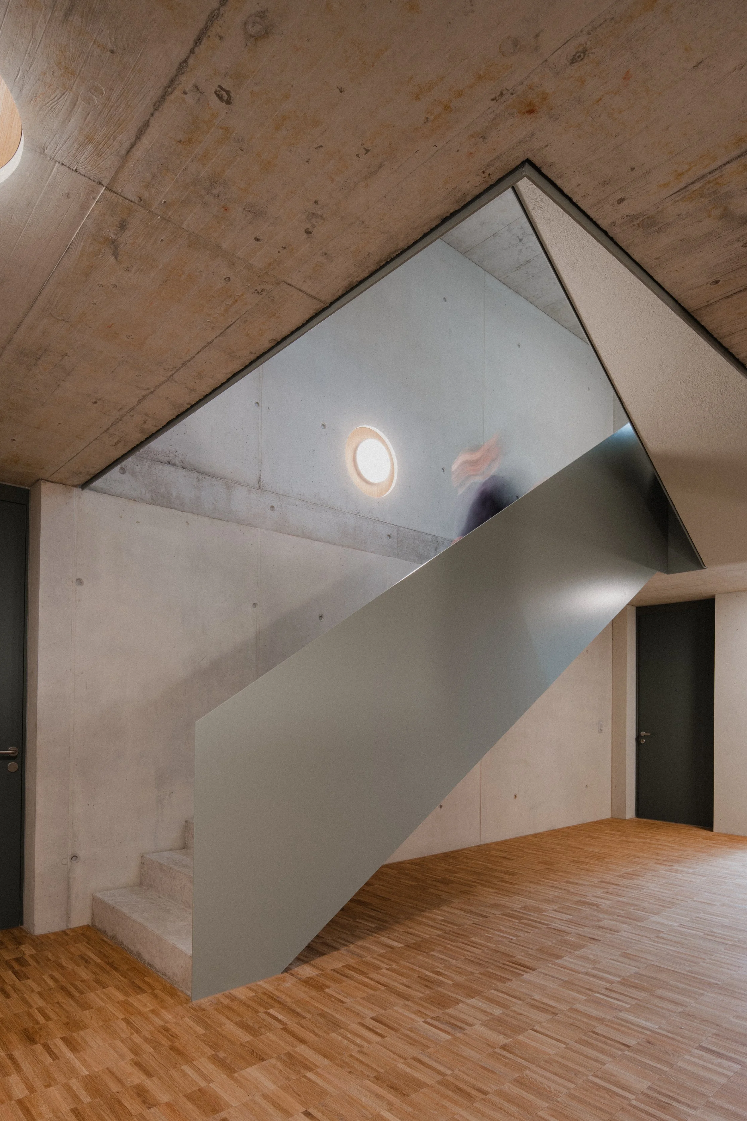 Innenaufnahme eines modernen Hauses mit Betonwänden, Holzdielenboden und einer markanten Treppe, Lichtquelle an der Decke, und Tür im Hintergrund.