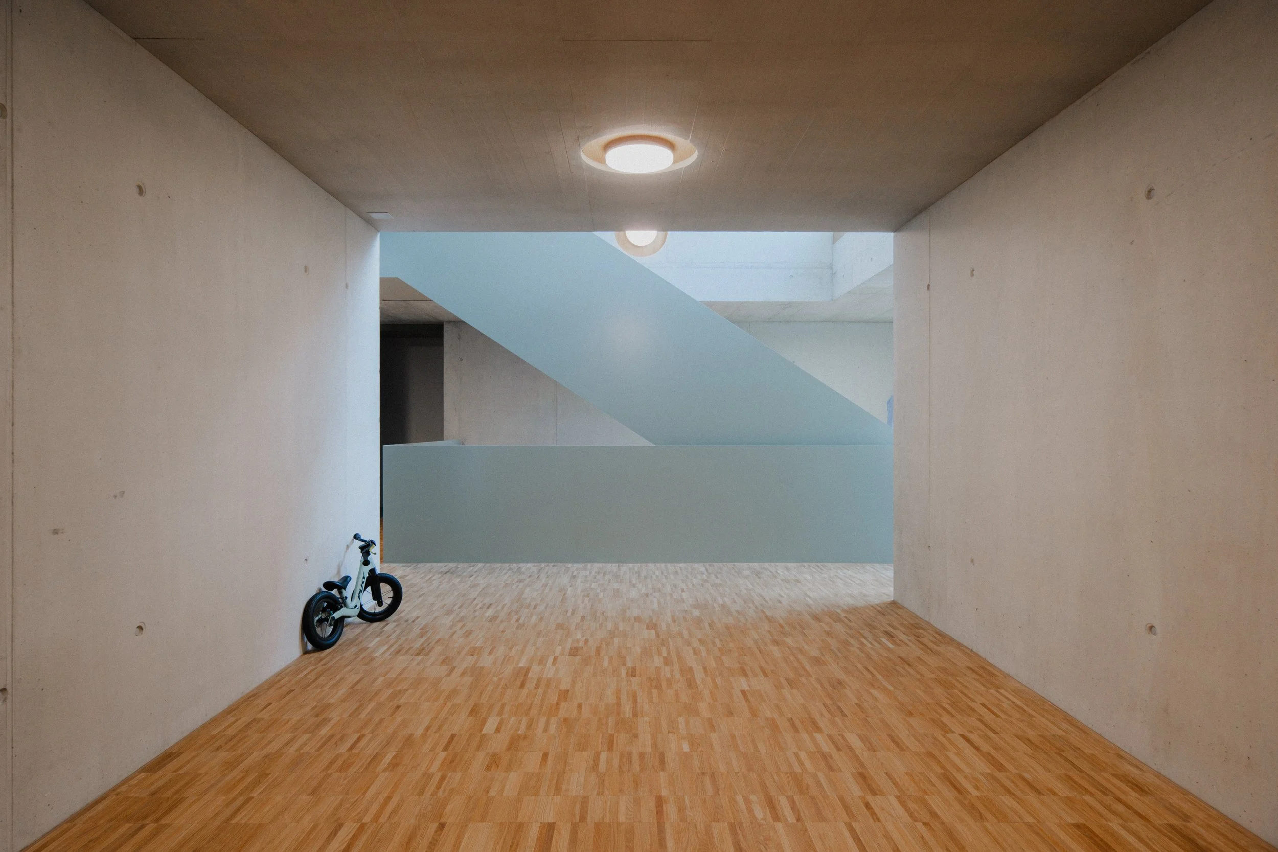 Innenaufnahme eines modernen Gebäudes mit weißen Wänden, Holzboden und einer Treppe im Hintergrund. Ein Kinderfahrrad steht an der Wand.