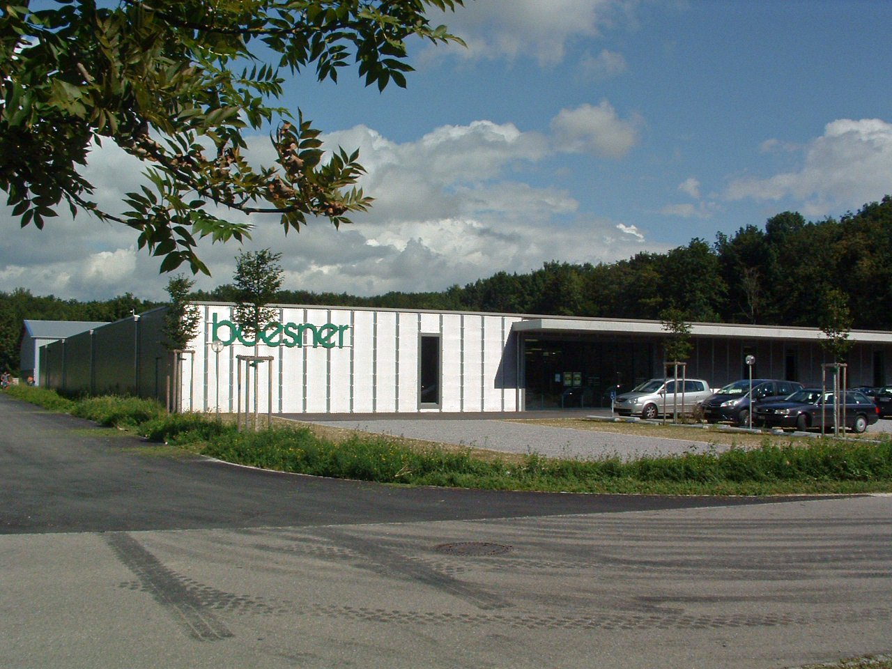 Ein modernes Gebäude mit dem Logo 'bosner' an der Seitenfassade, umgeben von Bäumen und einem Parkplatz mit Autos, bei sonnigem Himmel.