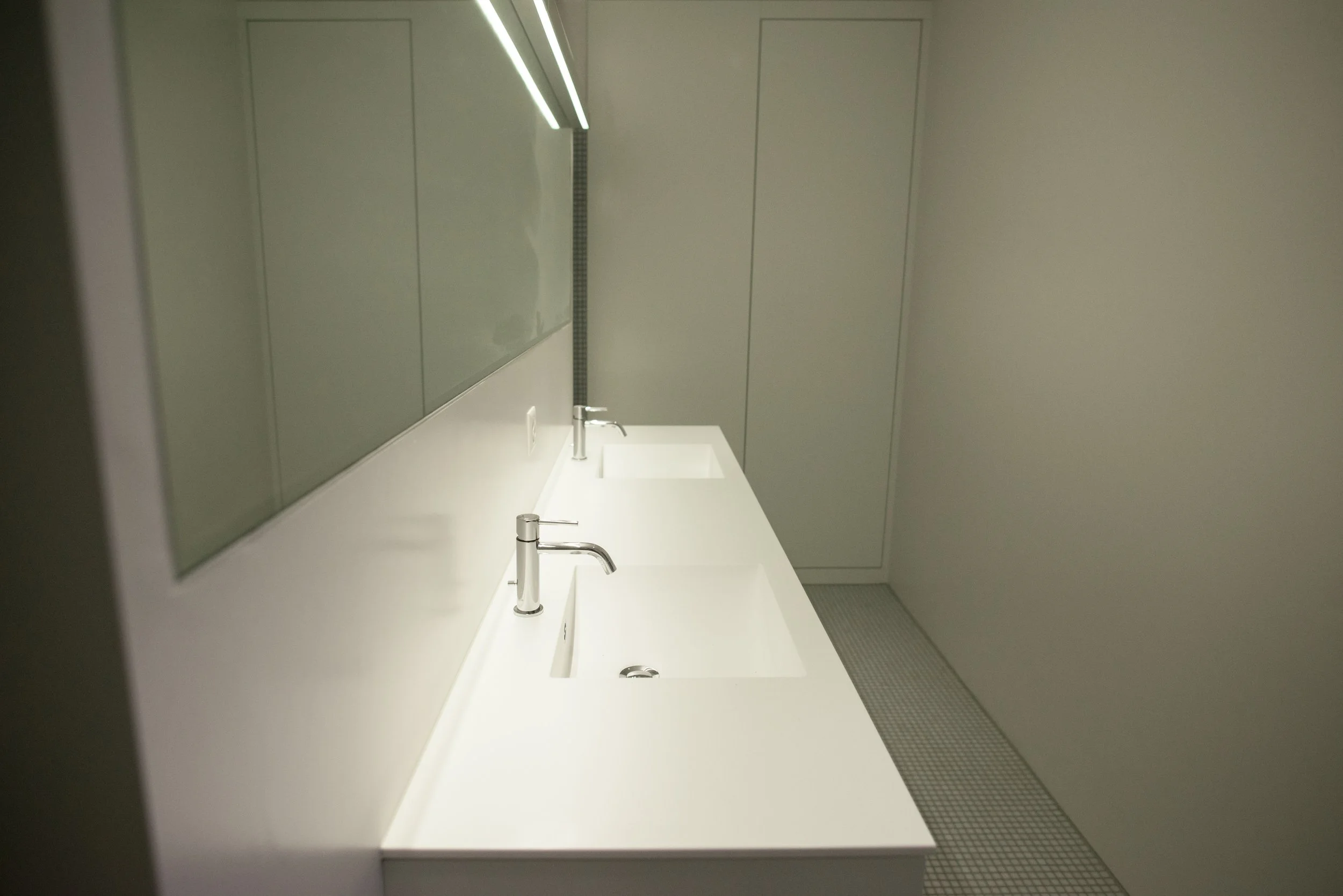 Ein moderner Waschraum mit weißen Waschbecken, jeweils mit einem Wasserhahn. Es gibt einen großen Spiegel an der Wand und den Boden aus kleinen Fliesen. Die Wände sind hell und schlicht gestaltet.