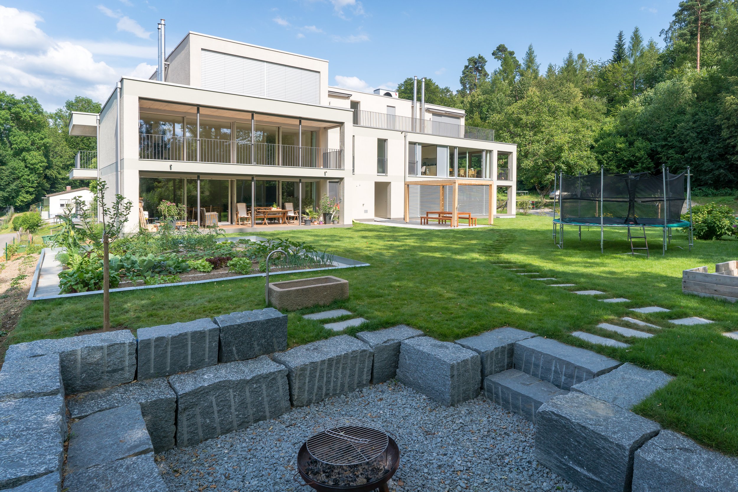 Ein moderner Mehrfamilienhaus mit Balkon und großen Fenstern, umgeben von einem gepflegten Garten mit Rasen, Blumen, einer Terrasse mit Sitzmöbeln, einem Trampolin im Hinterhof und einem Stein-Feuerstelle.