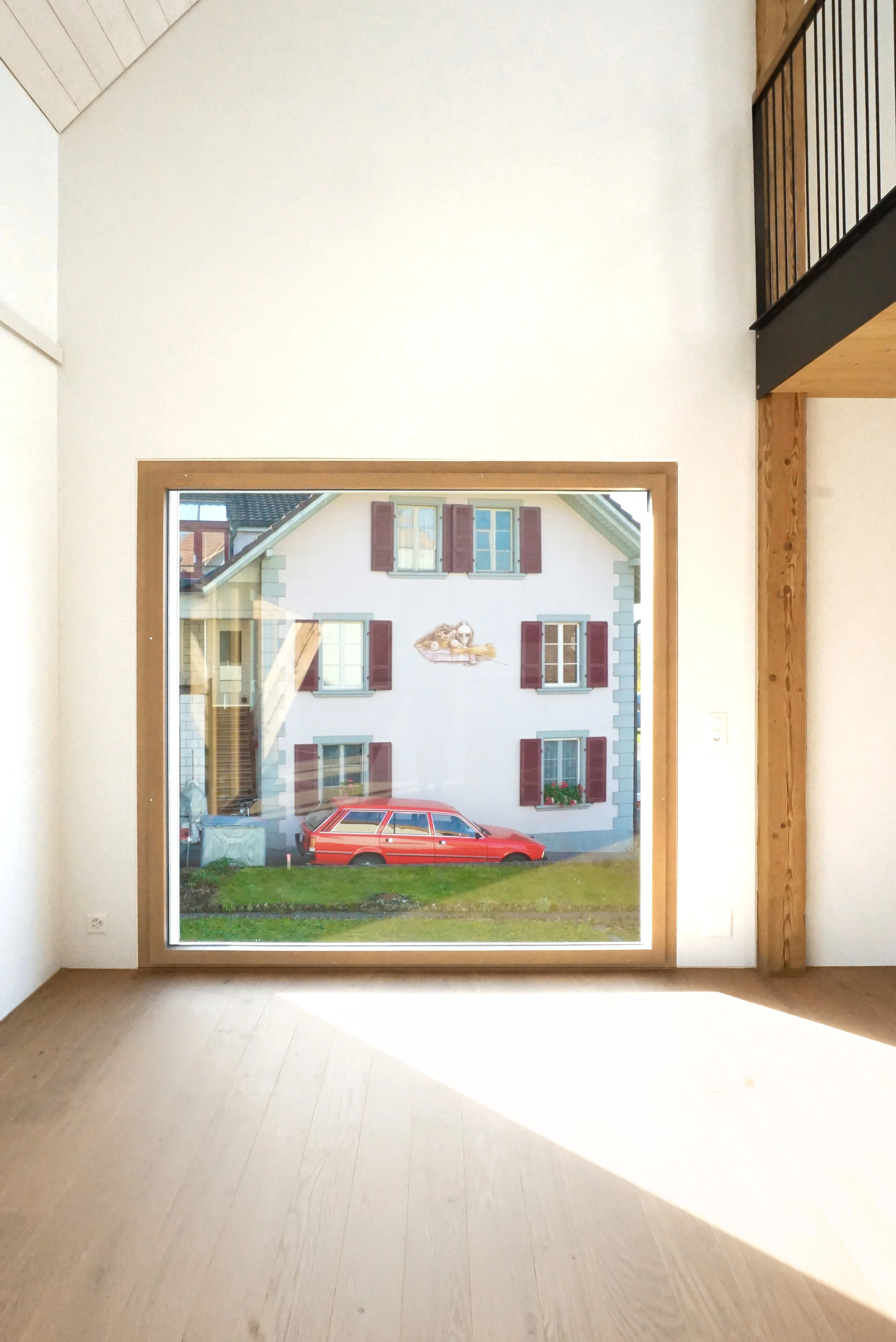 Ein großes Fenster zeigt das Äußere eines Hauses mit roten Fensterläden und einem roten Auto davor.