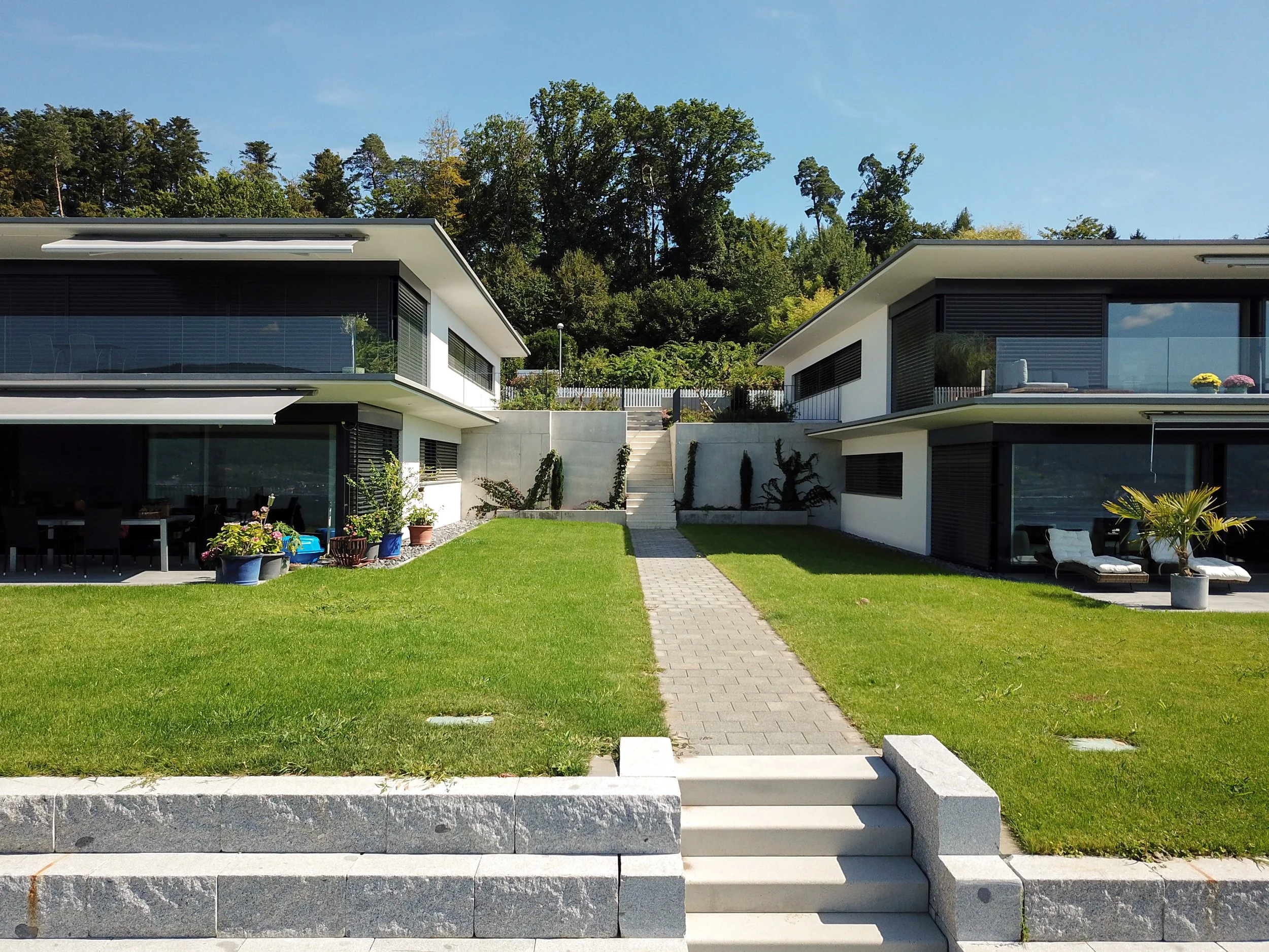Modernes Wohnhaus mit großen Glasfenstern, umgeben von einem gepflegten Garten, auf einer terrassierten Anhöhe mit Blick auf einen Waldrand, an einem sonnigen Tag.