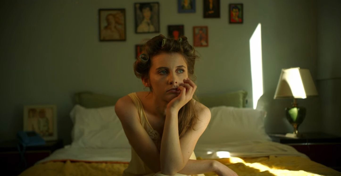 A woman with hair rollers sitting on a bed in a bedroom, looking thoughtful, with sunlight coming through a closed door and framed artwork on the wall behind her.