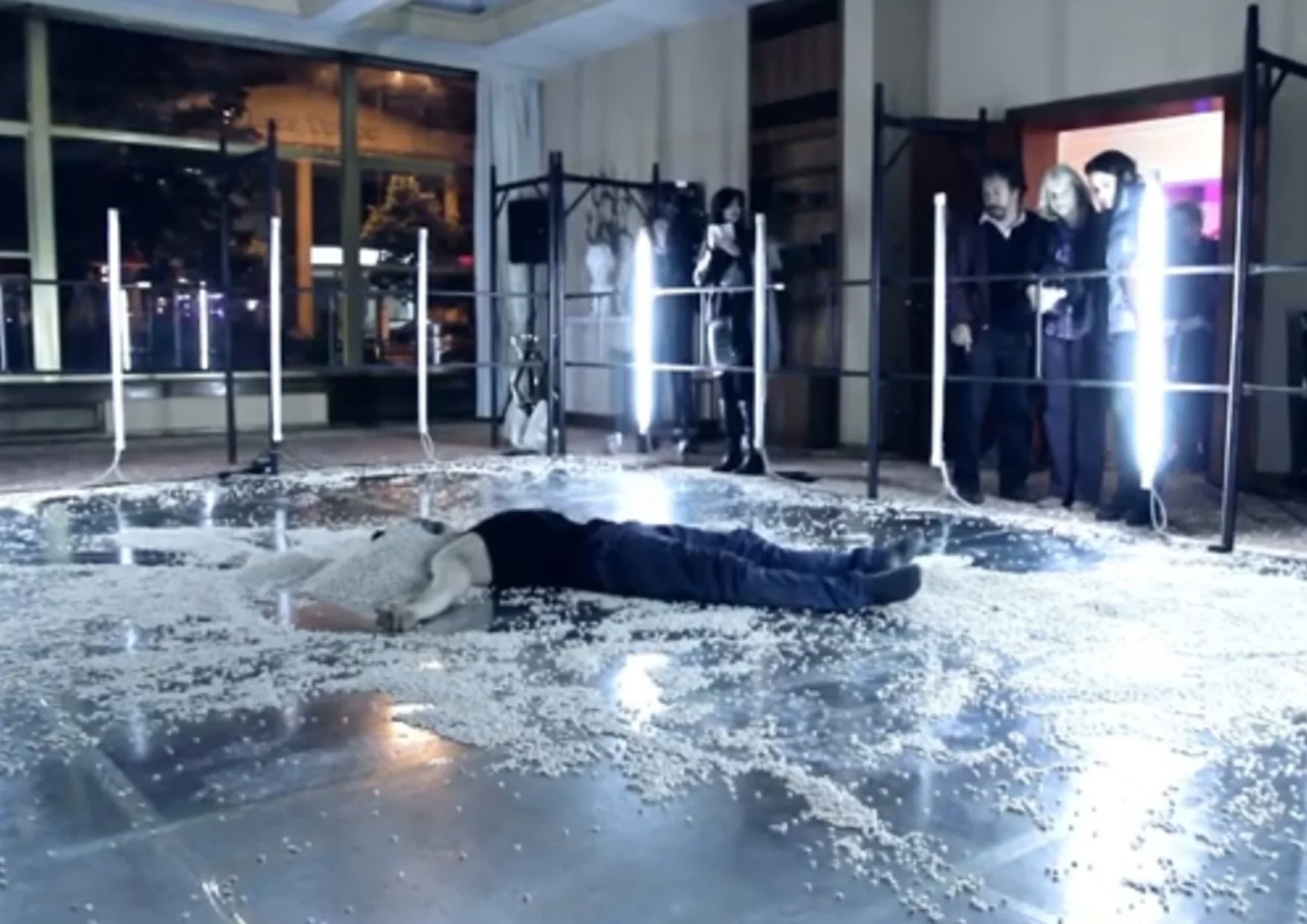 Person lying face down on the floor covered with scattered rice in a room with large windows, and several people observing through a railing.