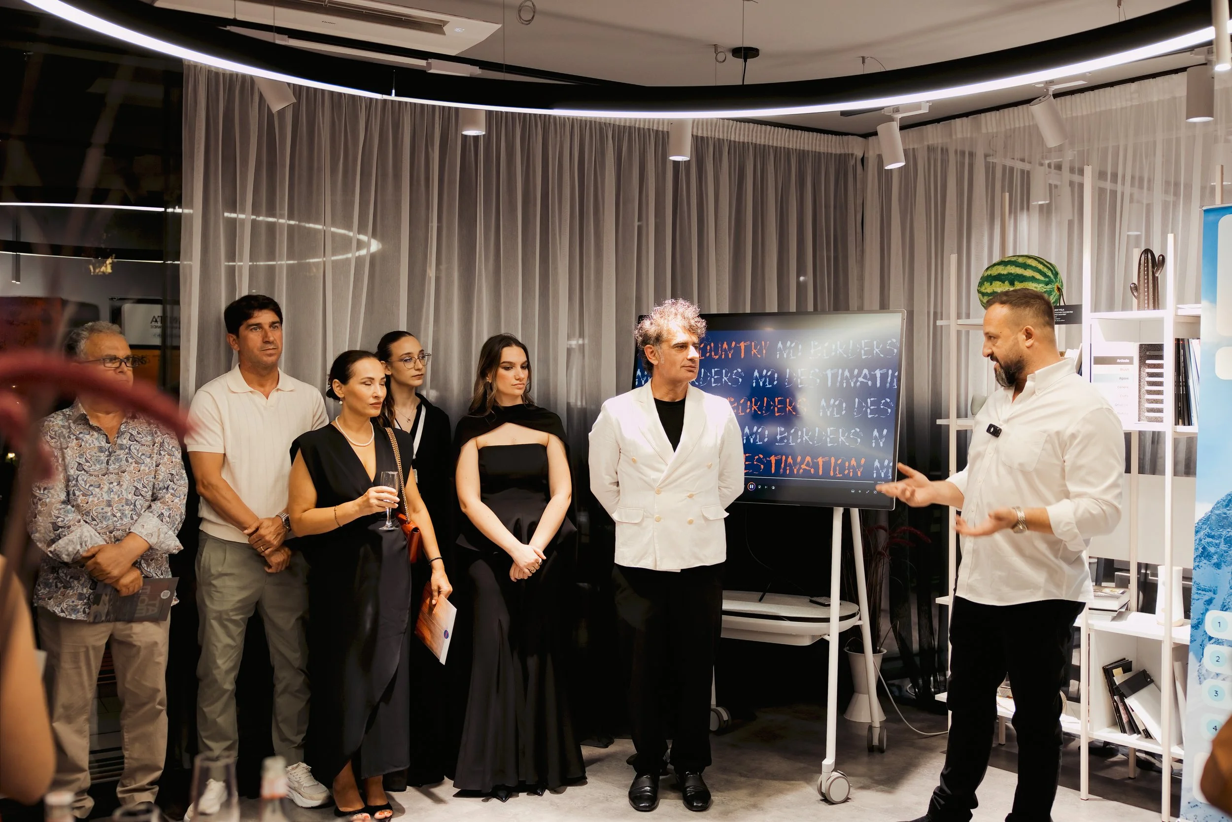 Group of six people standing and listening to a man in white shirt, who appears to be explaining something. They are indoors, with a large digital screen behind them displaying text, and decorative items like a watermelon and cacti on shelves.