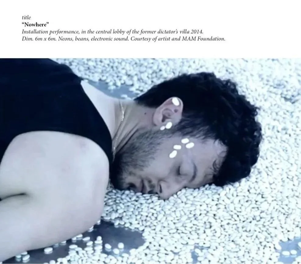 A person lying on the floor with their cheek on a large pile of white beans or small white objects, with white dots on their face and ear, dressed in a black sleeveless top.