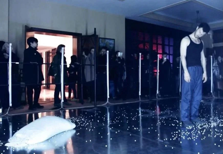 An art installation with a mannequin in casual clothes standing on a shiny floor sprinkled with small lights, with a group of people observing behind a barrier in a dark room with red and blue lighting.