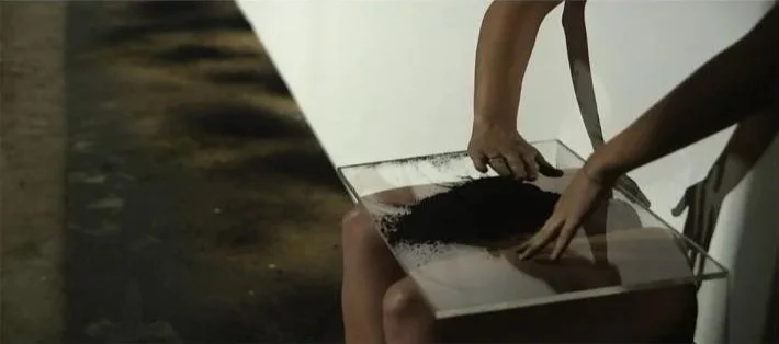 Two people handling black powdery substance over a glass surface, with water nearby.