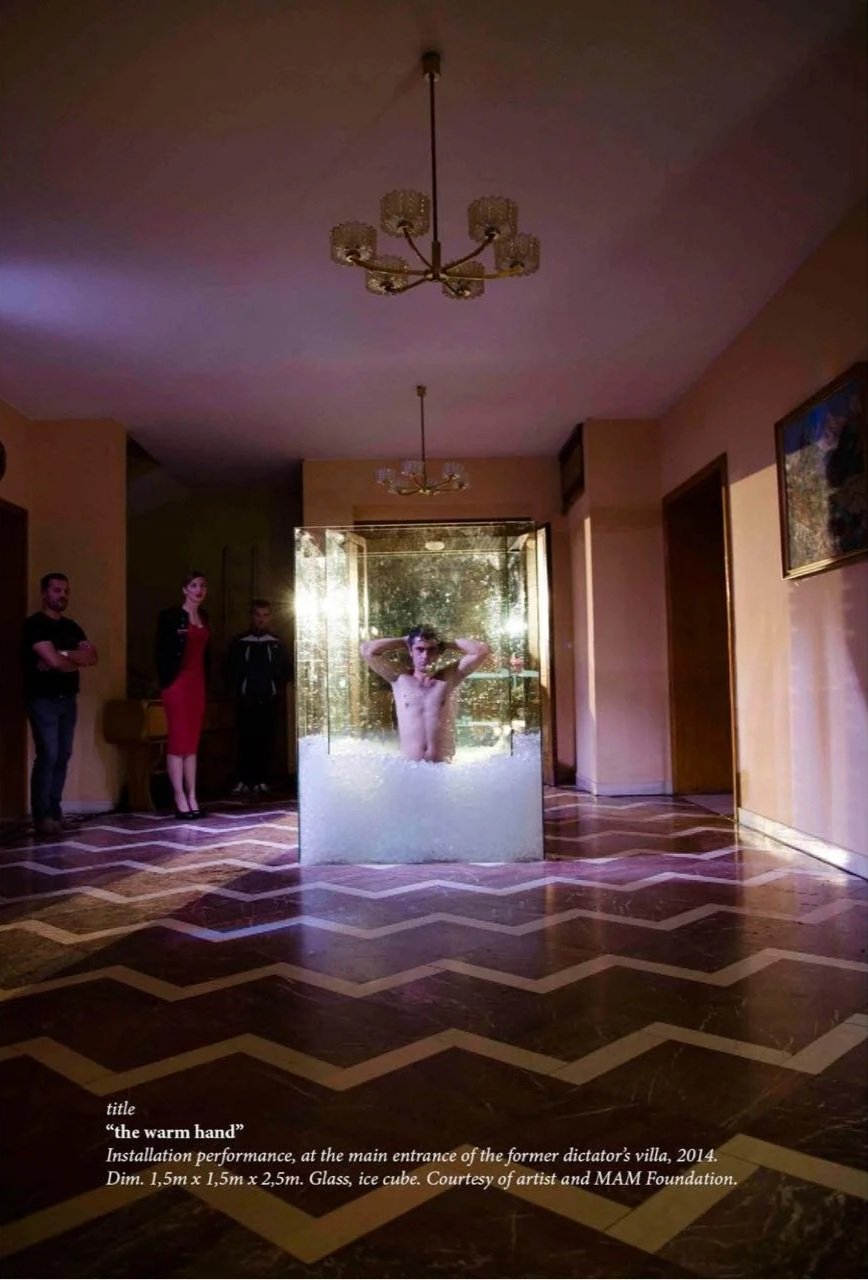 An indoor art installation shows a man submerged in a glass ice cube with his hands behind his head, located at the main entrance of a former dictator's villa. Three people stand nearby, observing the piece, with the room decorated with chandeliers a