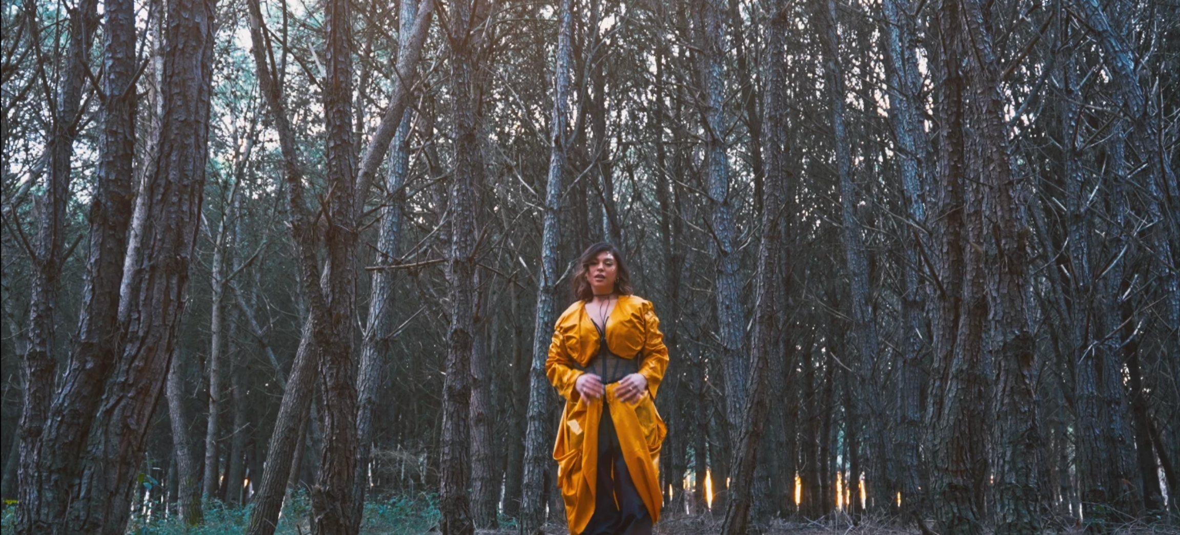 A woman in a yellow jacket and black clothing standing in a dense forest of tall, dark trees with light filtering through the branches.