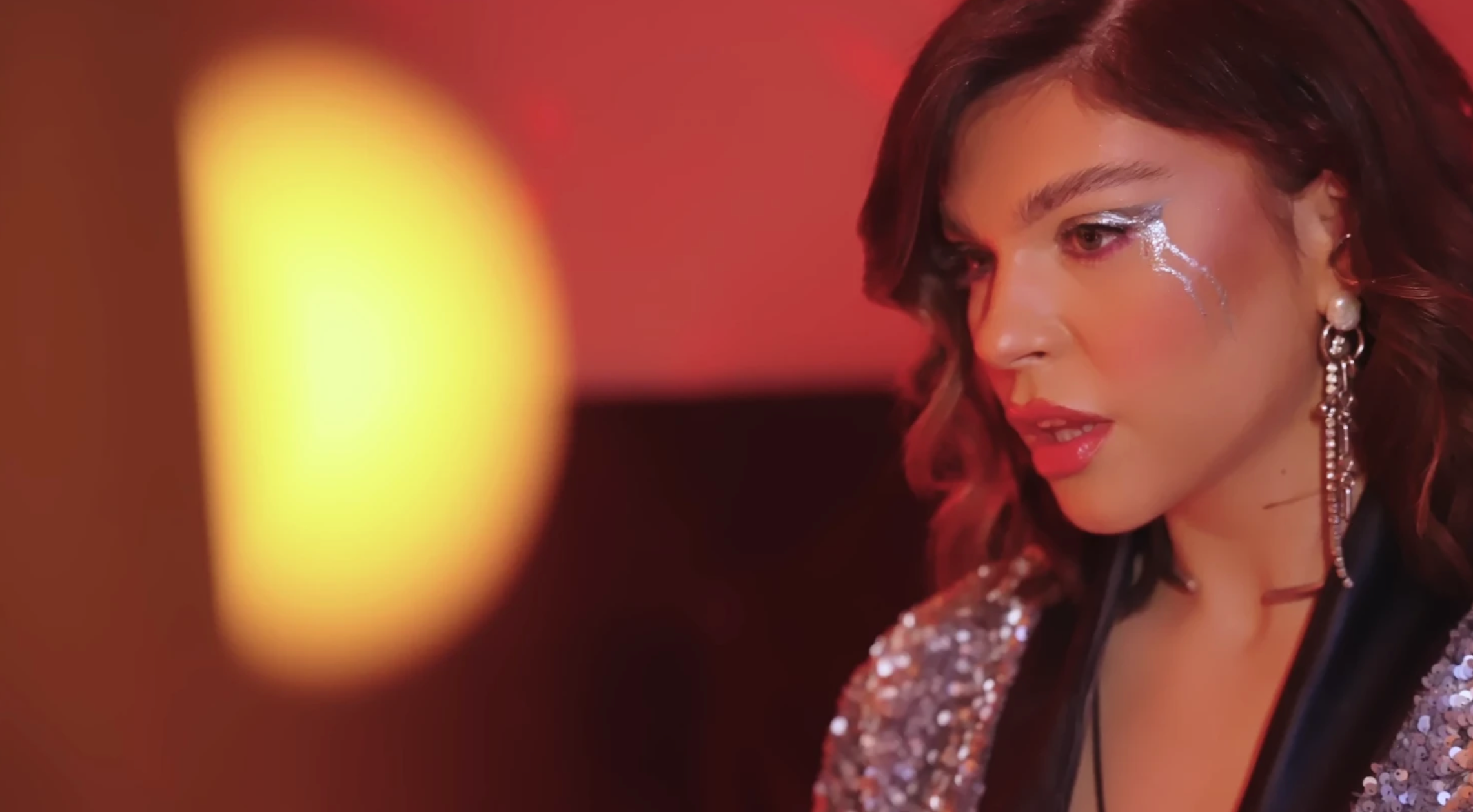 A woman with wavy hair and glamorous makeup, including glittery silver eyeshadow, wearing large earrings and a sequined top, looking to the side against a warm, blurred background.
