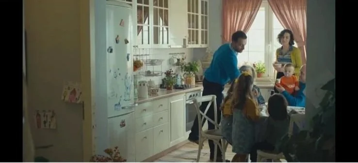 Family gathered around a kitchen table with a man serving food, children, and a woman standing near the window.