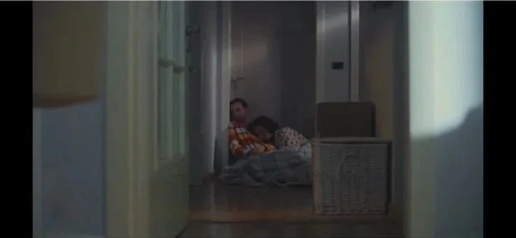 A person and a child sitting on the floor in a hallway near a washing machine and refrigerator, with a basket nearby.