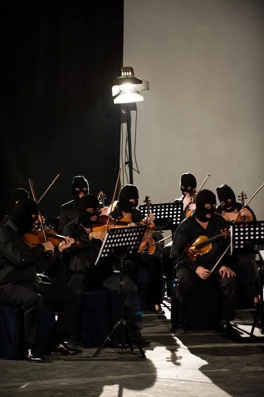 Orchestra of musicians wearing black masks and suits, playing violins and other string instruments on stage with music stands, dimly lit with a spotlight from above.