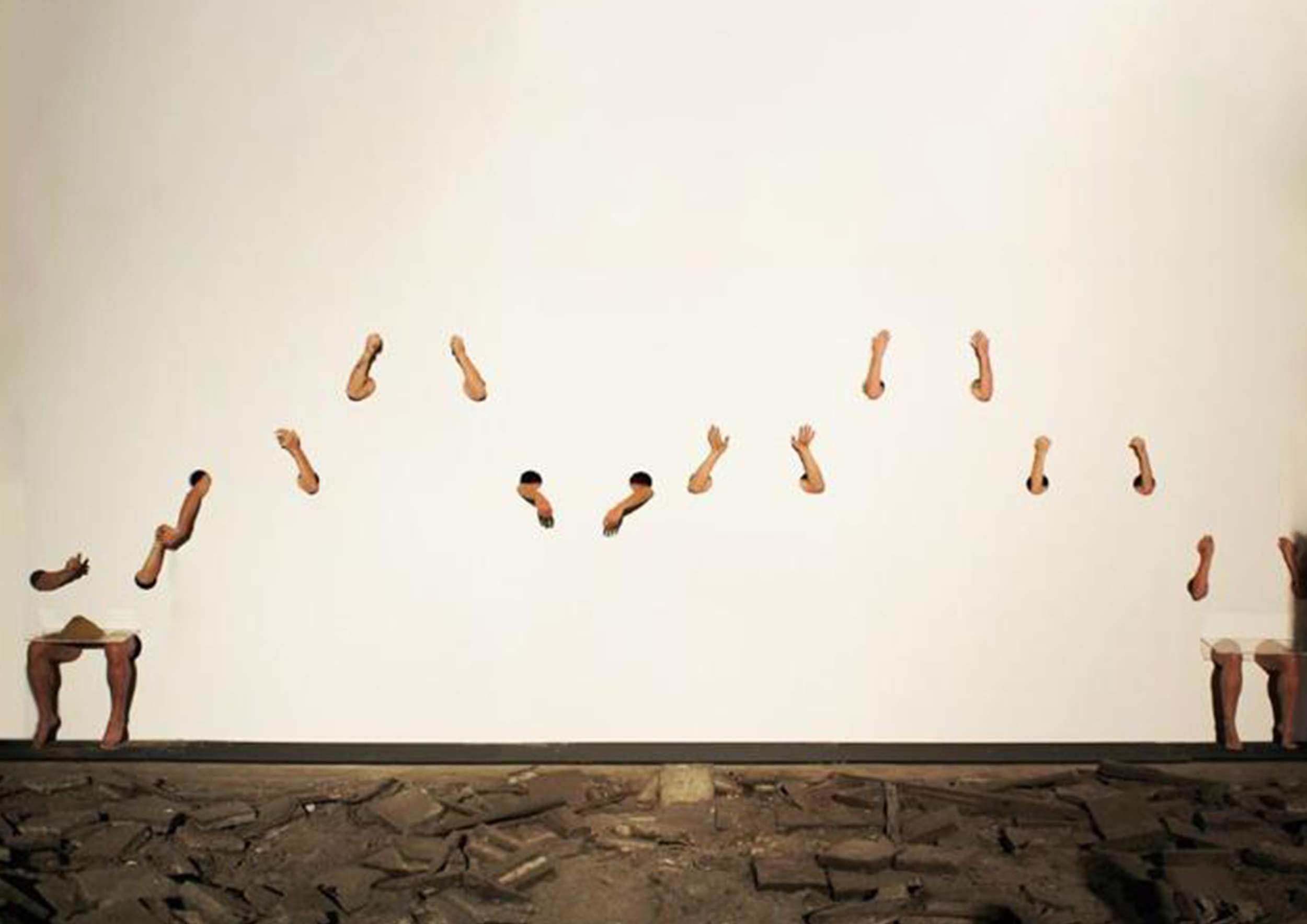 A performance artwork with human arms and legs protruding from the wall, arranged in a pattern, with a person in a suit at the bottom left corner.