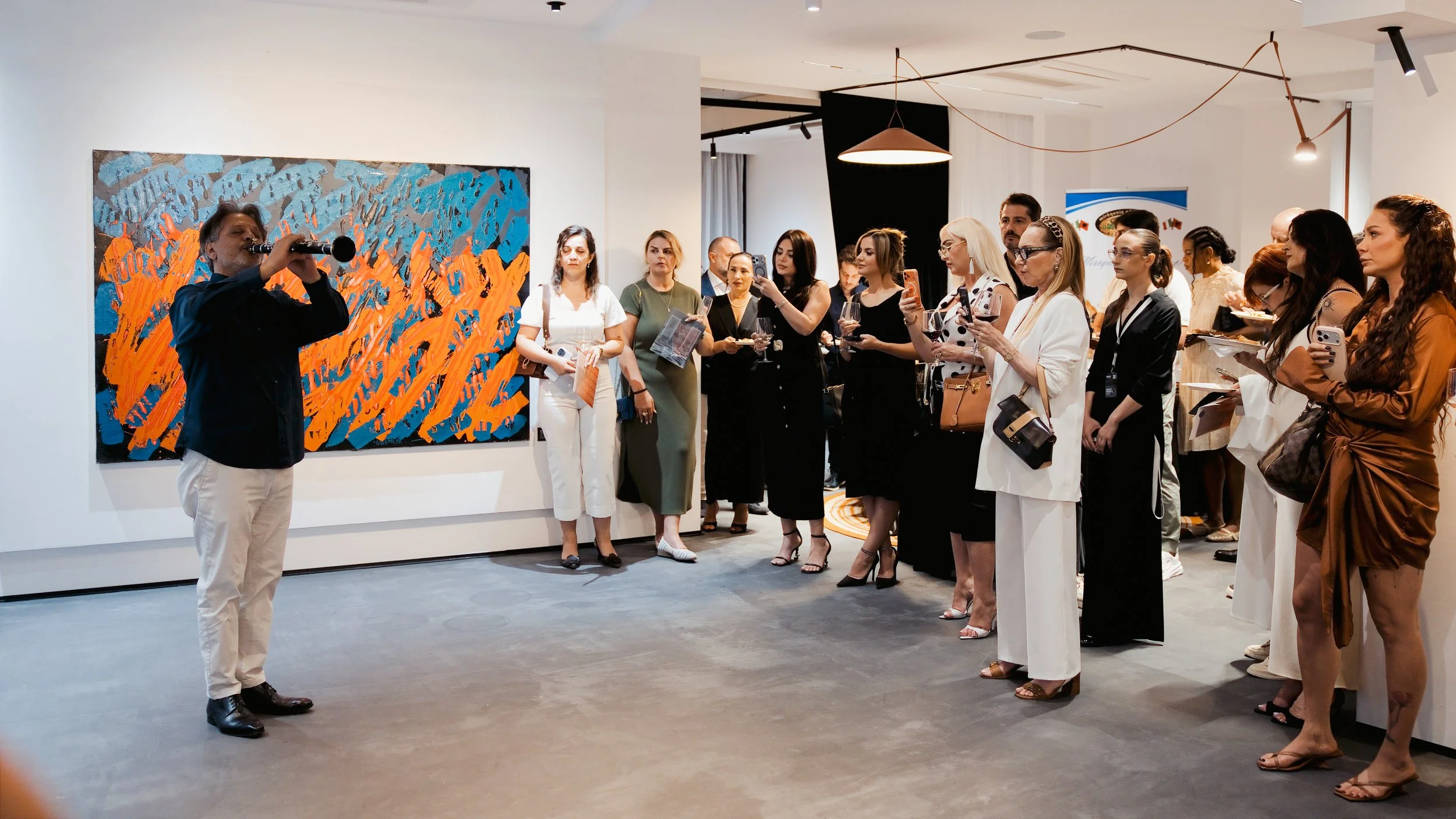Group of people attending an art exhibition, listening to a person playing a wind instrument, with colorful abstract painting on the wall.