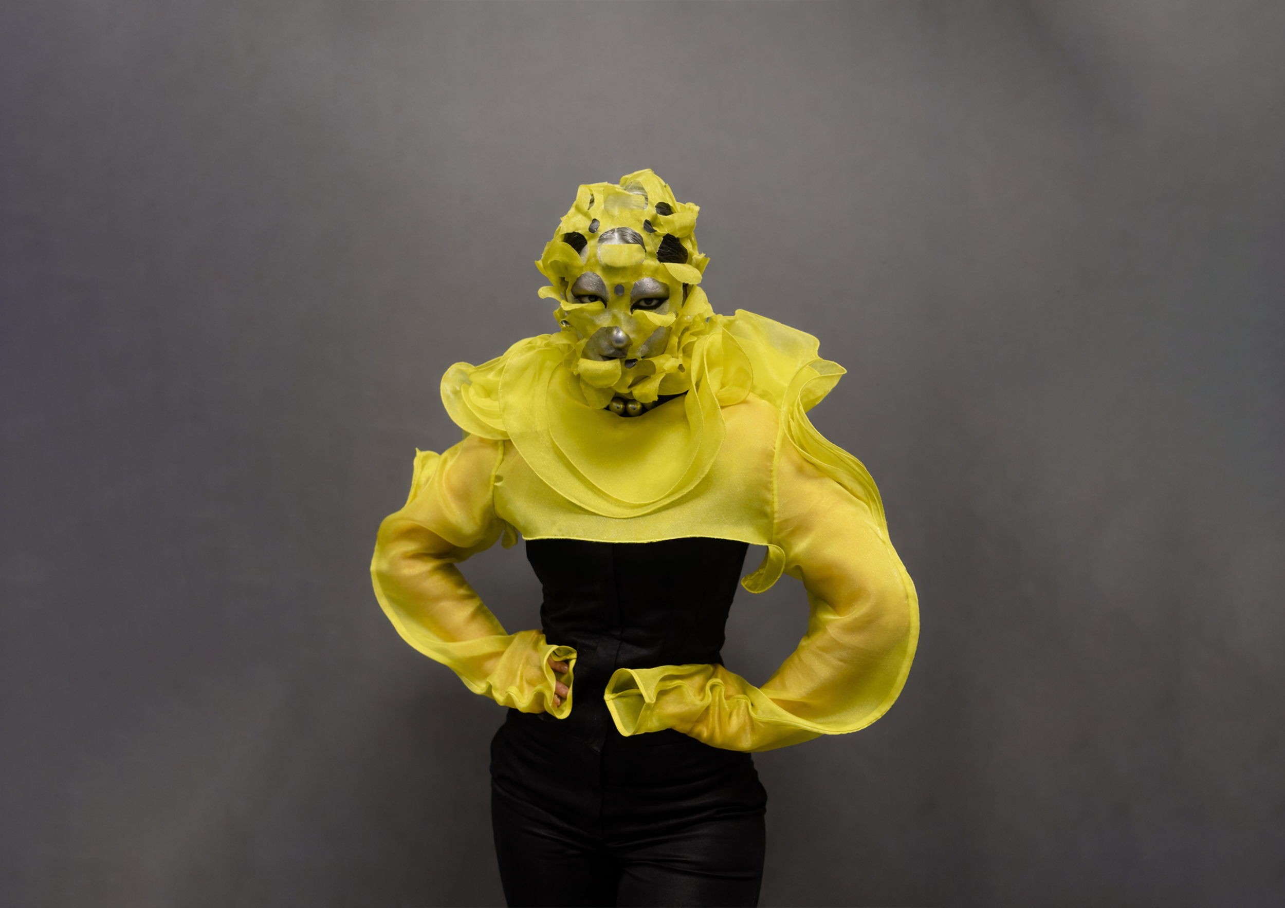A person in a yellow ruffled jacket with a mask resembling a dog's face, posing with one hand on their hip against a plain gray background.