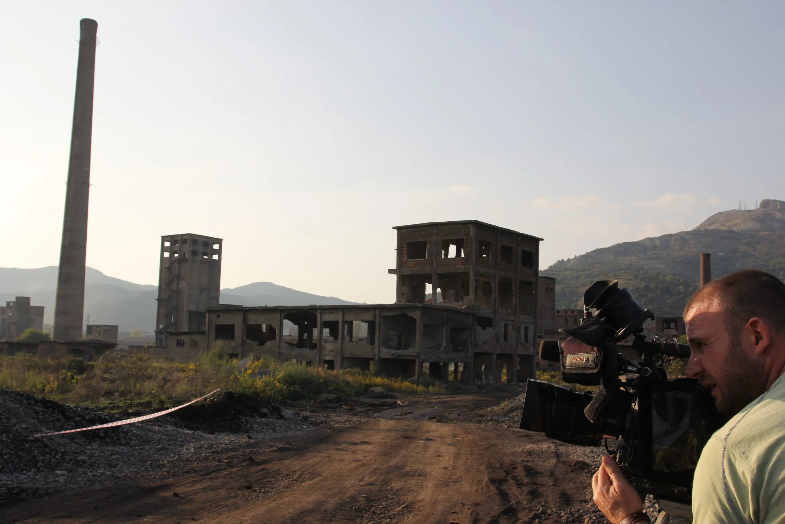 Person using a professional camera to film or photograph an abandoned industrial building with mountains in the background.