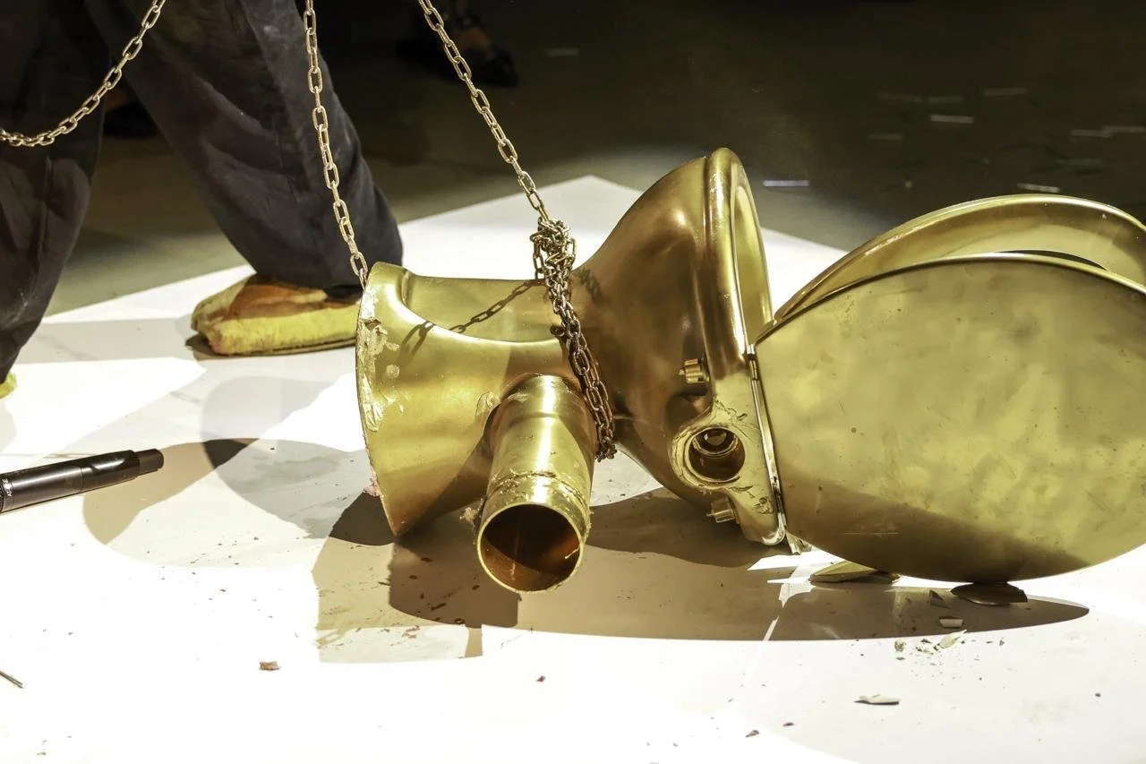A broken brass boat propeller chained to a stand, lying on a white surface with a person standing nearby.