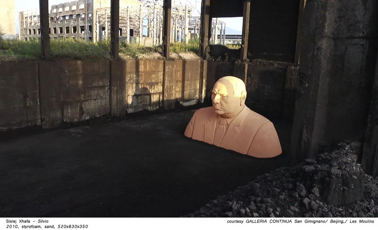 A large beige bust sculpture of a man in a suit, positioned on the ground in a dark, unfinished space with visible concrete walls and supports. Outside, there is a building with multiple windows and some greenery.