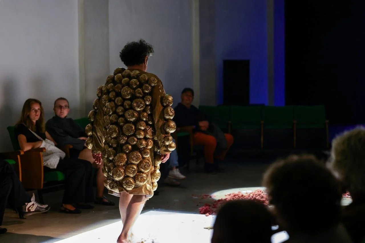 A person walking in a room wearing a coat covered with metallic or shiny round objects, possibly resembling buttons or decorative elements, while people seated watch.
