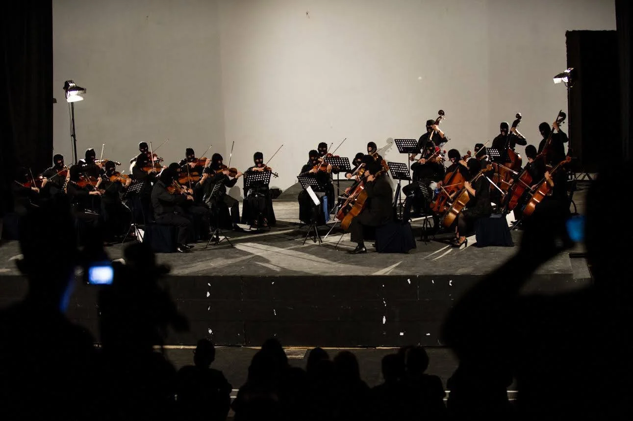 Orchestra of musicians playing string instruments on stage, with audience members taking photos in the dark.