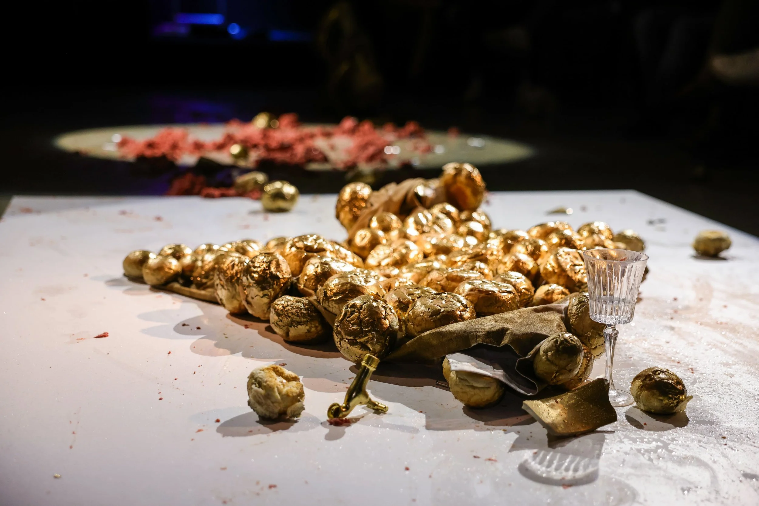 Golden foil-wrapped chocolate eggs scattered on a white table with some fallen pieces around, a small clear glass nearby, and a dark background with blurred items.