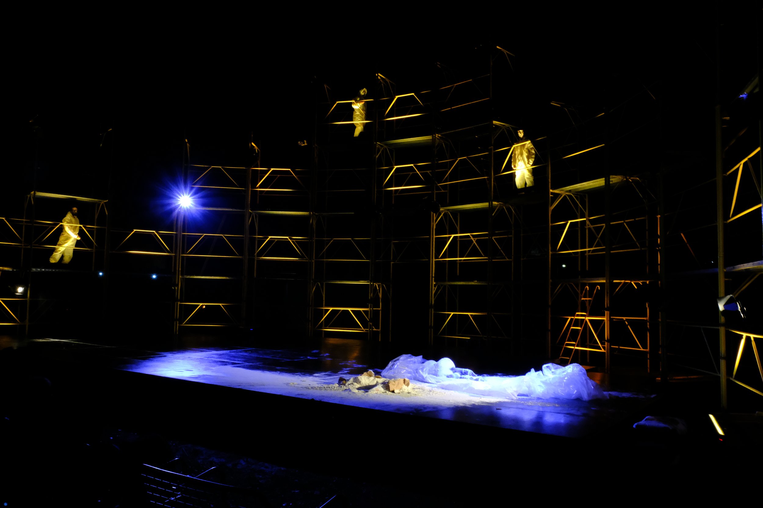 Nighttime scene of a multilevel scaffolding stage with performers in gold costumes illuminated by yellow lighting, blue light on stage, and some stage props on the ground.