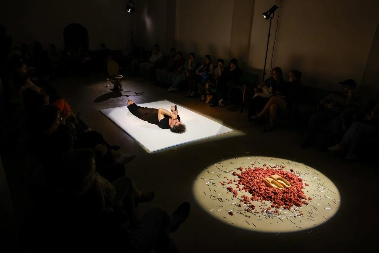 Person lying on their side on a lit white platform, holding a microphone, in a dark room with an audience seated around. A circular spotlight on the floor features red material with two objects and scattered matches.